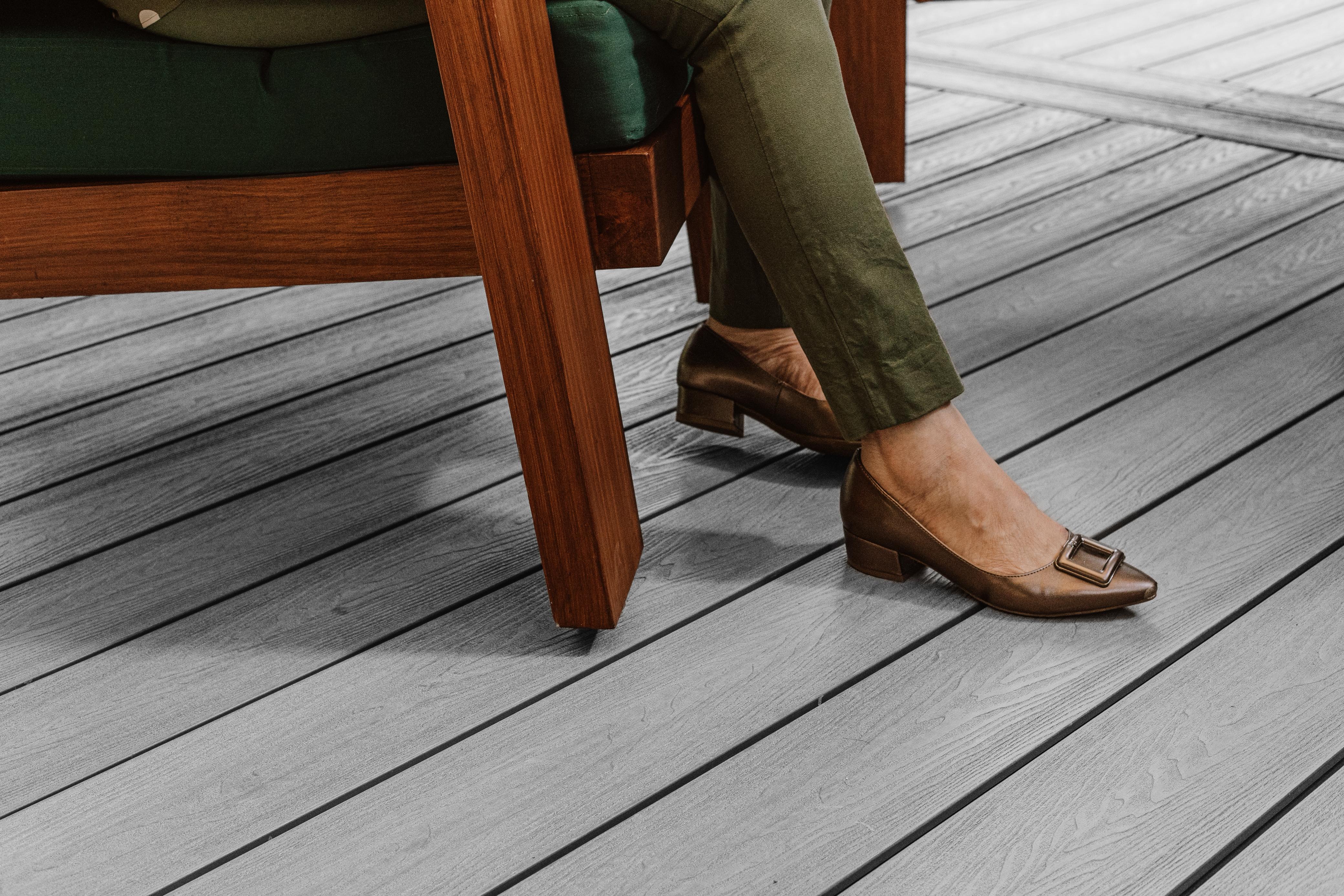 Piernas con pantalones verdes y zapatos marrones con tacón bajo, bajo una silla de madera.