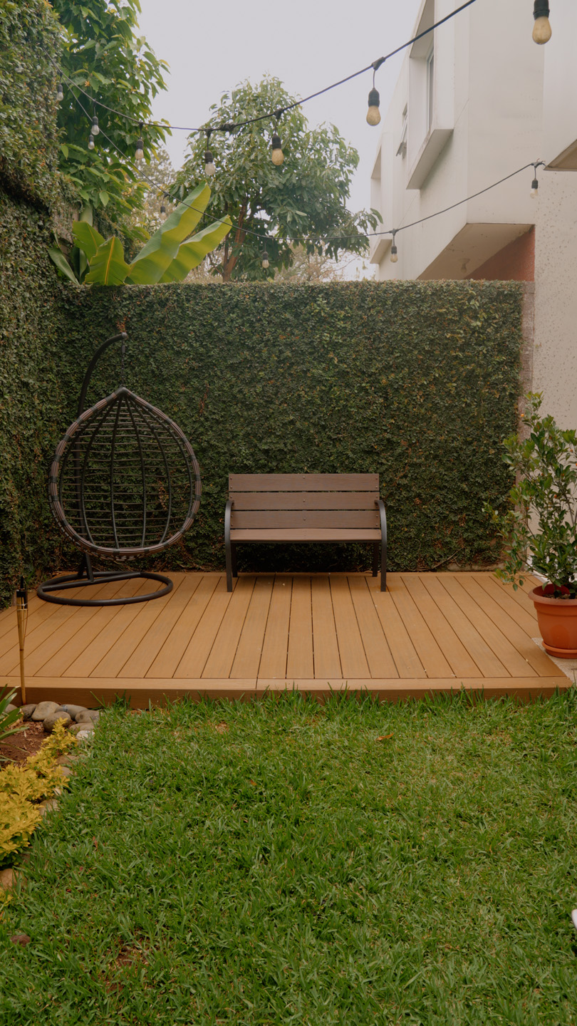 Patio con suelo de madera, banco, silla colgante y plantas verdes en jardín pequeño.