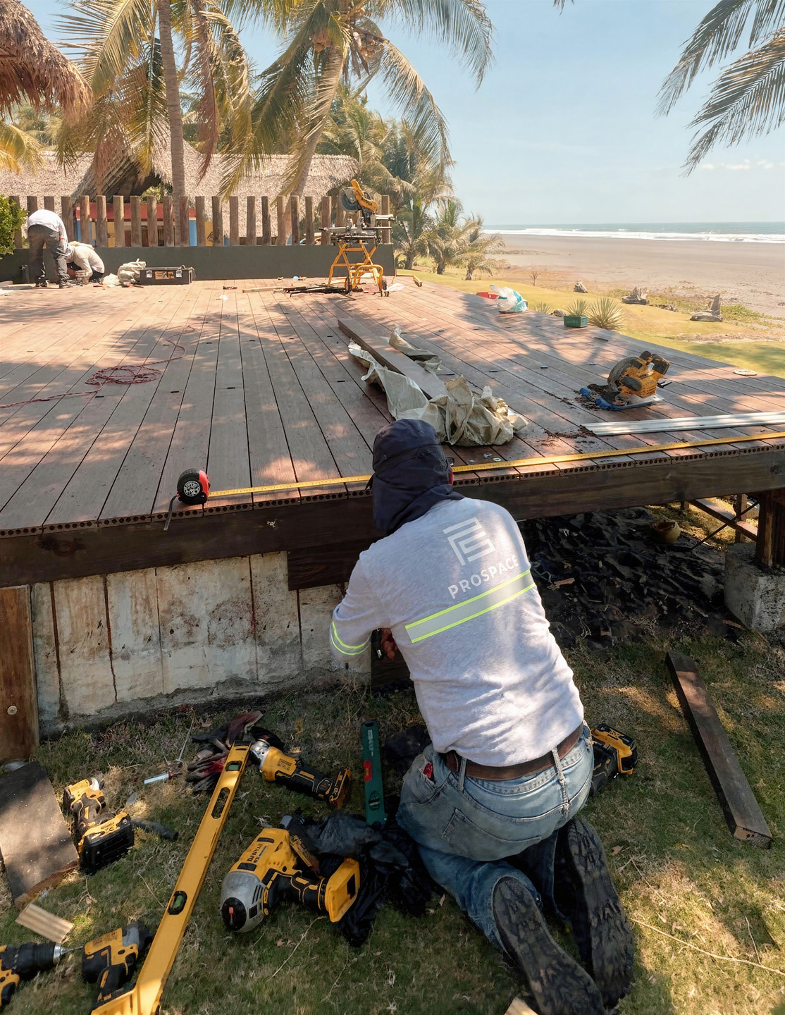 Trabajador arrodillado realiza construcción de plataforma de madera cerca de playa con palmeras.