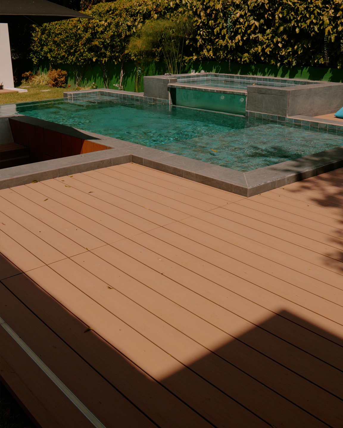 Piscina con mosaicos azules y un jacuzzi elevado en un área exterior con piso de madera y plantas.