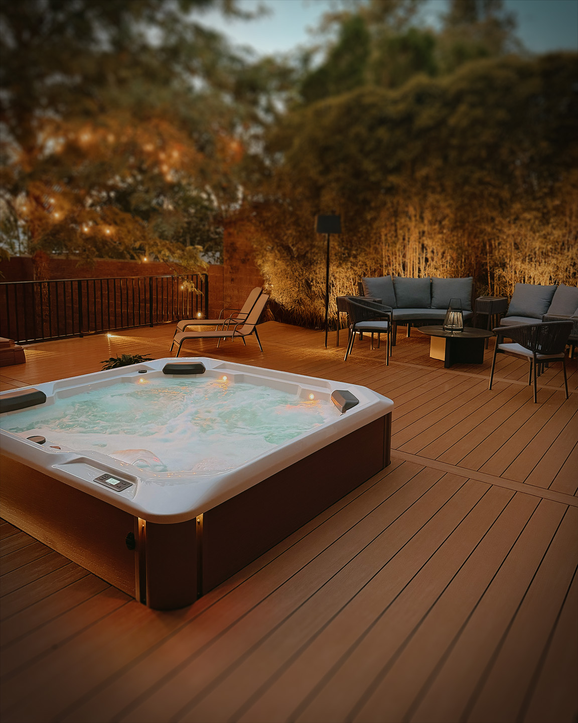 Jacuzzi encendido en terraza de madera con muebles de exterior y luces cálidas al atardecer.