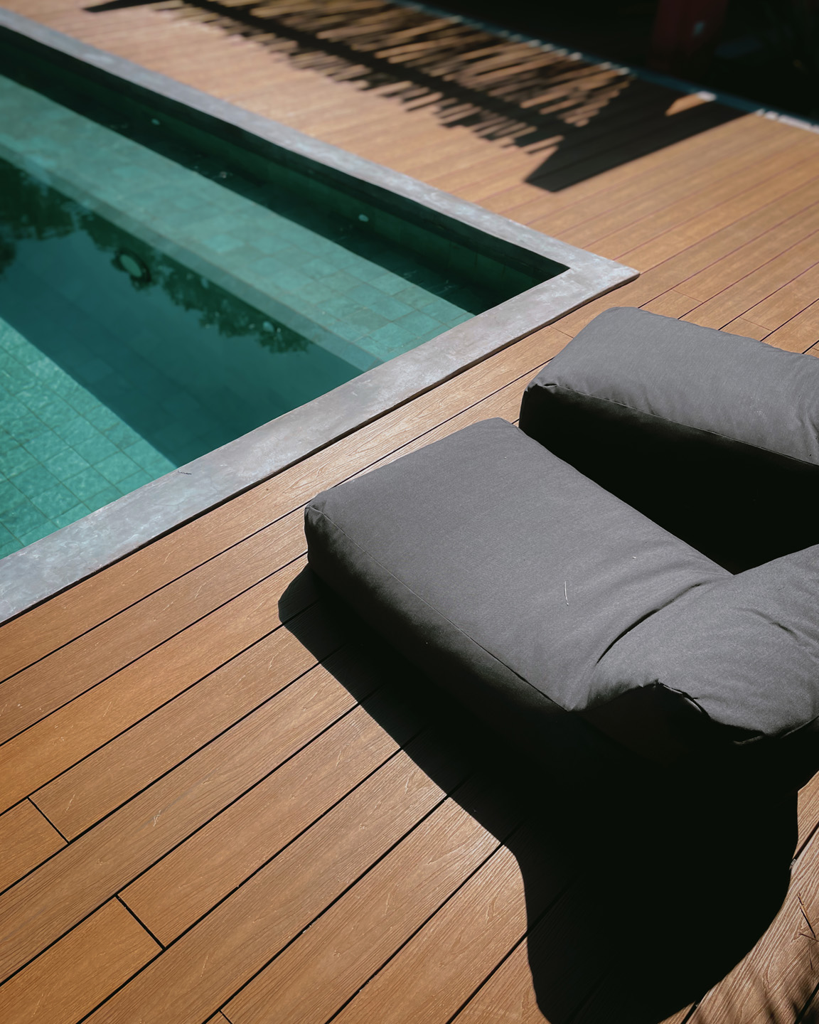 Almohadón gris grande sobre piso de madera junto a piscina con azulejos verdes.