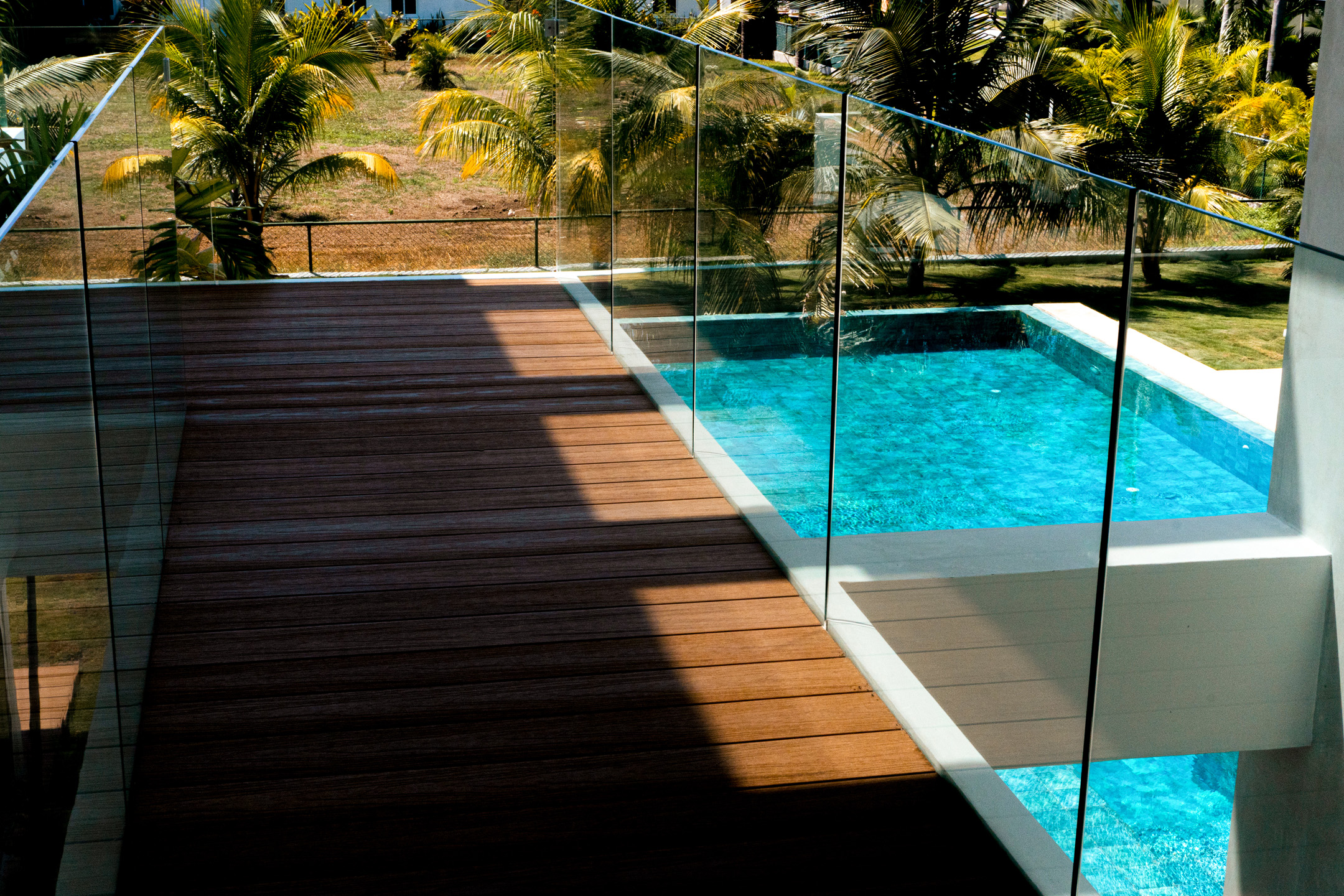 Terraza con piso de madera y piscina con agua azul rodeada de palmeras.