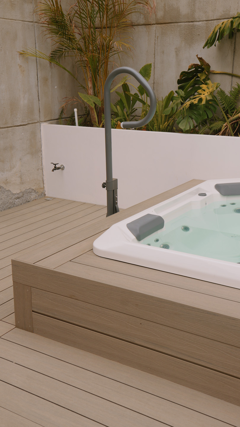 Jacuzzi al aire libre con plataforma de madera y plantas verdes a su alrededor.
