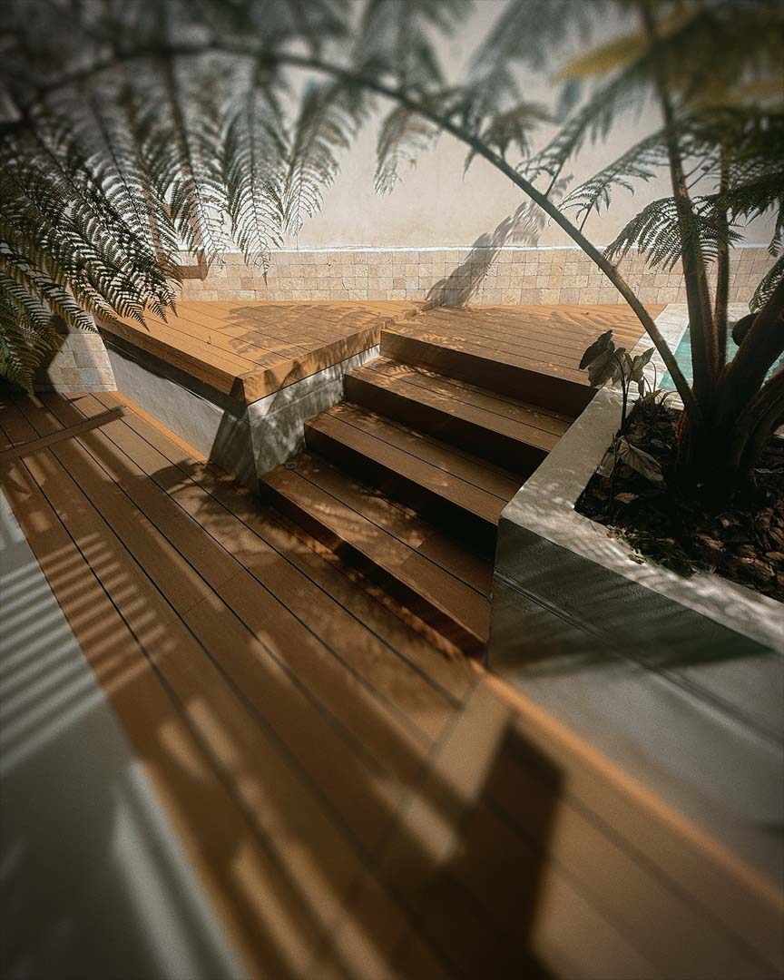 Escaleras y plataforma de madera junto a planta en maceta con pared de azulejos beige.