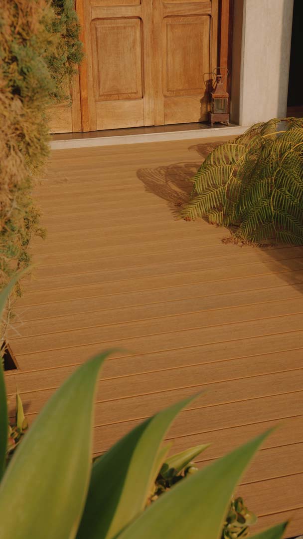 Entrada con puerta de madera, piso de tablas y plantas verdes a los lados.