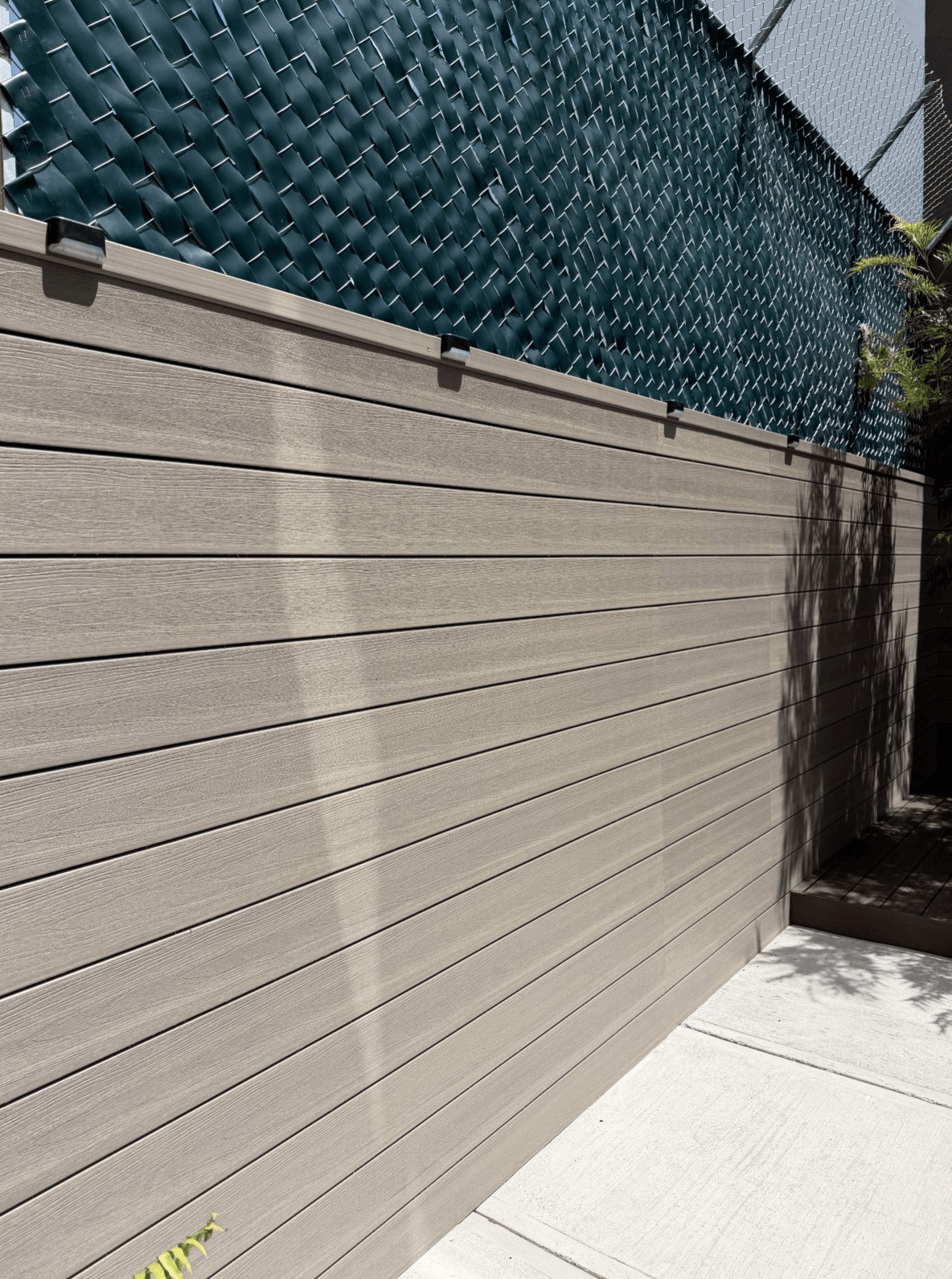 Cerca de madera gris con malla metálica azul y suelo de concreto bajo luz solar.