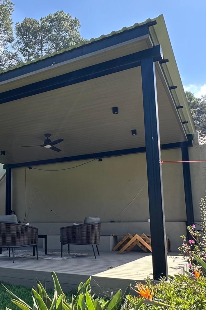 Área exterior con pérgola negra, sillas de mimbre, ventilador de techo y plantas a la orilla.