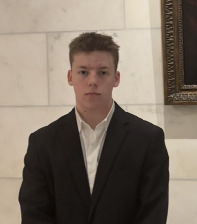 Young man with short hair wearing a black suit and white shirt standing against a marble wall.