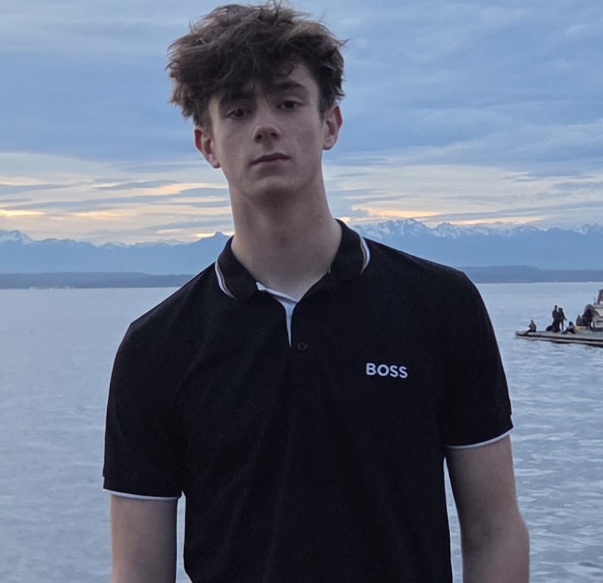 Young man wearing black BOSS polo shirt standing by a lake with mountains and cloudy sky in the background.