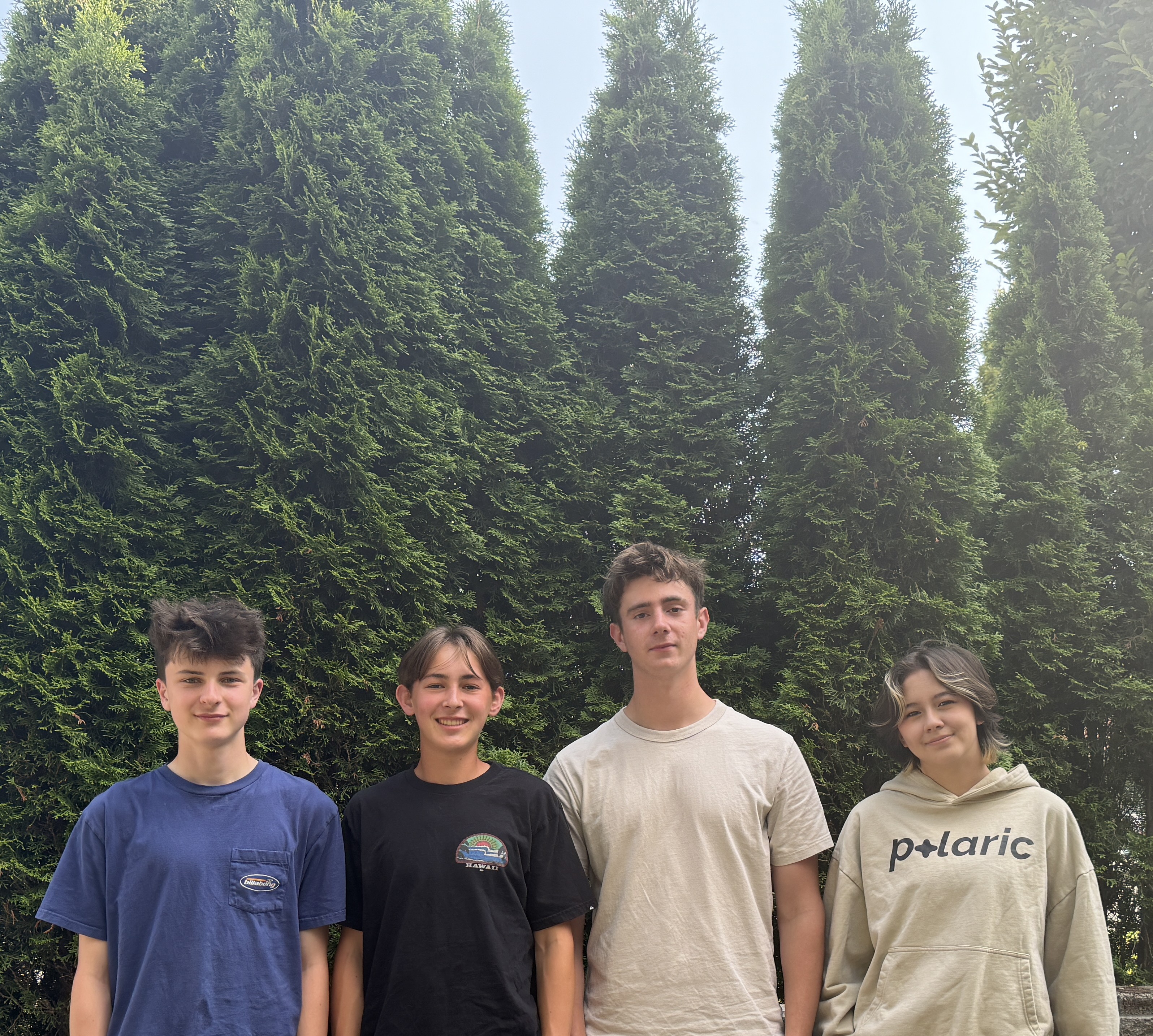 Four teenagers standing side by side outdoors in front of tall green trees, smiling at the camera.