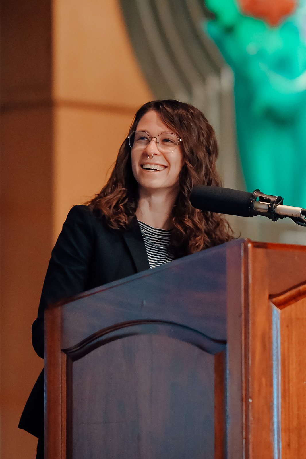 Keynote speaker in glasses laughing at a wooden podium with a teal glass sculpture behind.