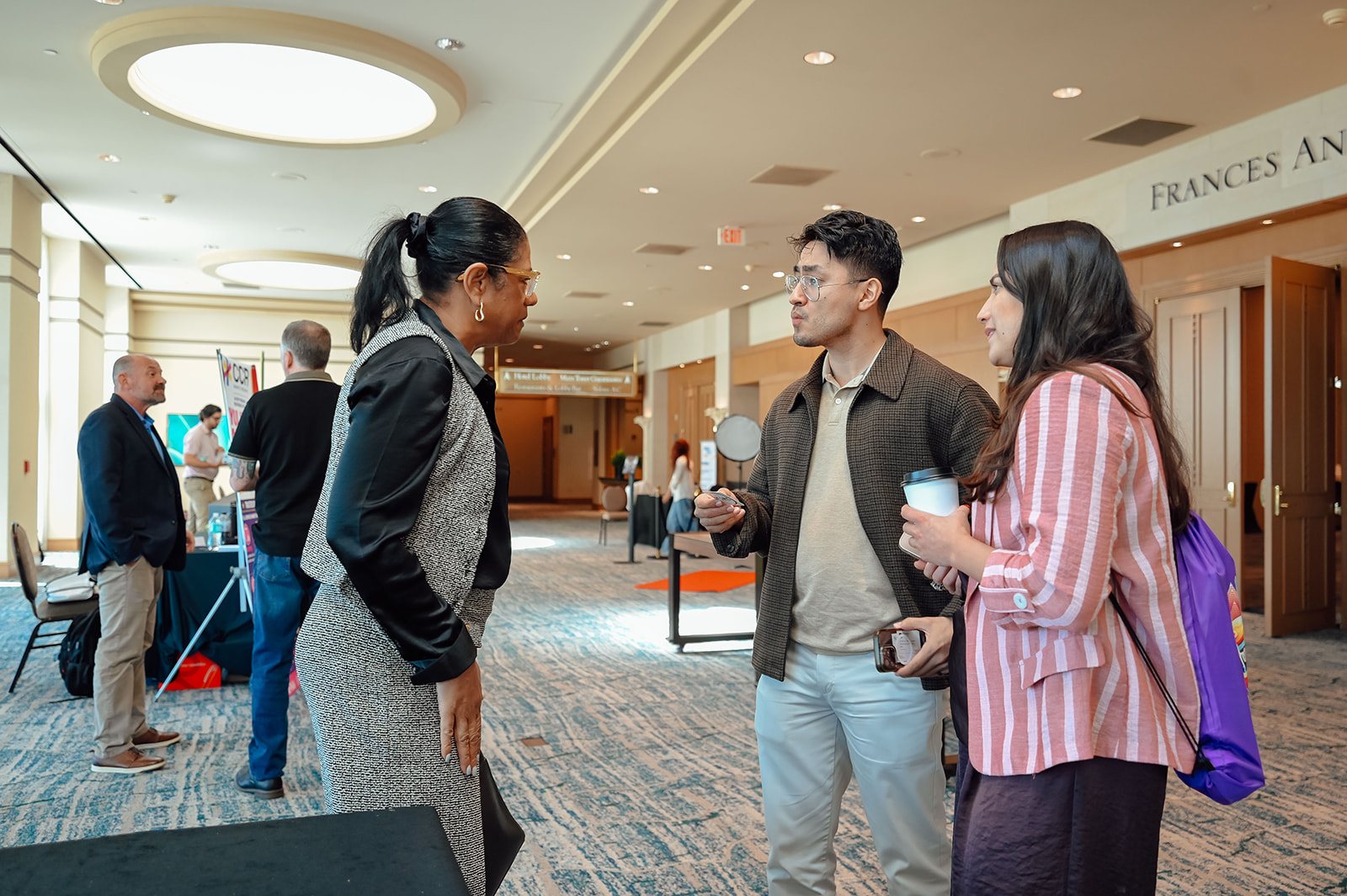 Three attendees chatting in a carpeted pre-function corridor with exhibitor tables behind.