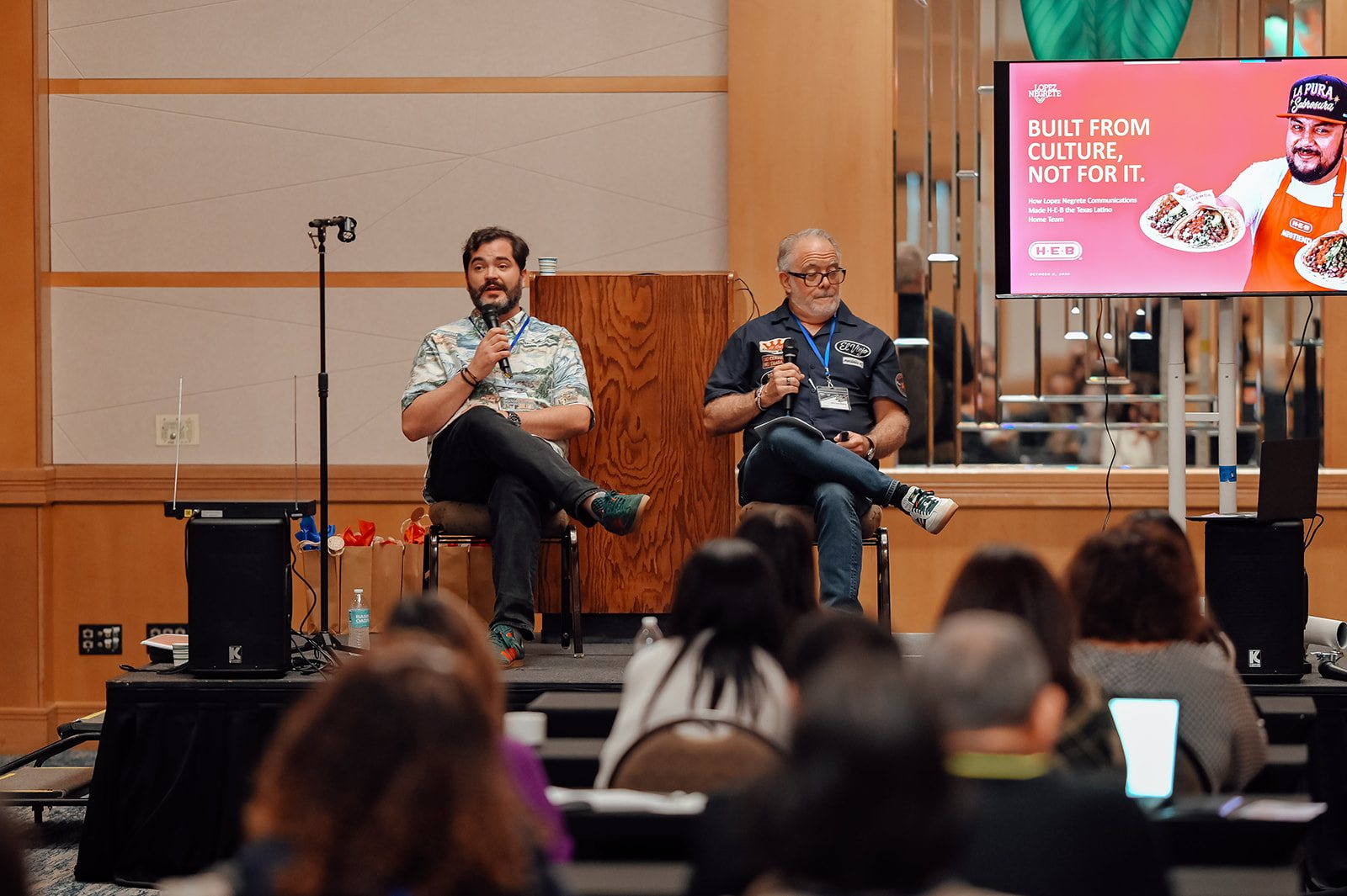 Two presenters seated on a low stage with handheld mics beside a ‘Built from Culture, Not for It’ slide with the Lopez Negrete and H-E-B logos.