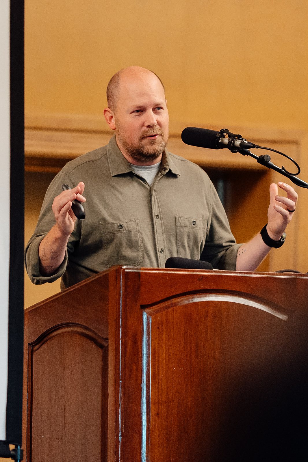 Speaker in an olive button-up shirt gesturing with a slide clicker at a wooden lectern.