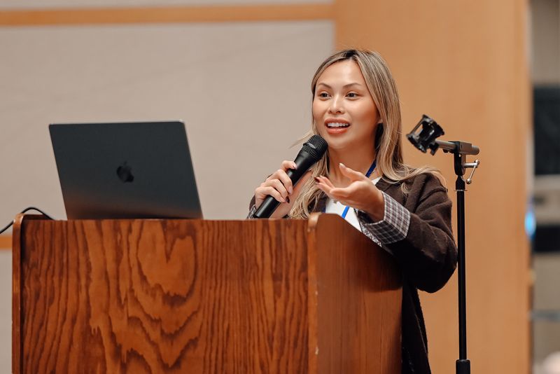 Presenter at a wooden lectern with a laptop, smiling and gesturing while speaking into a handheld mic.