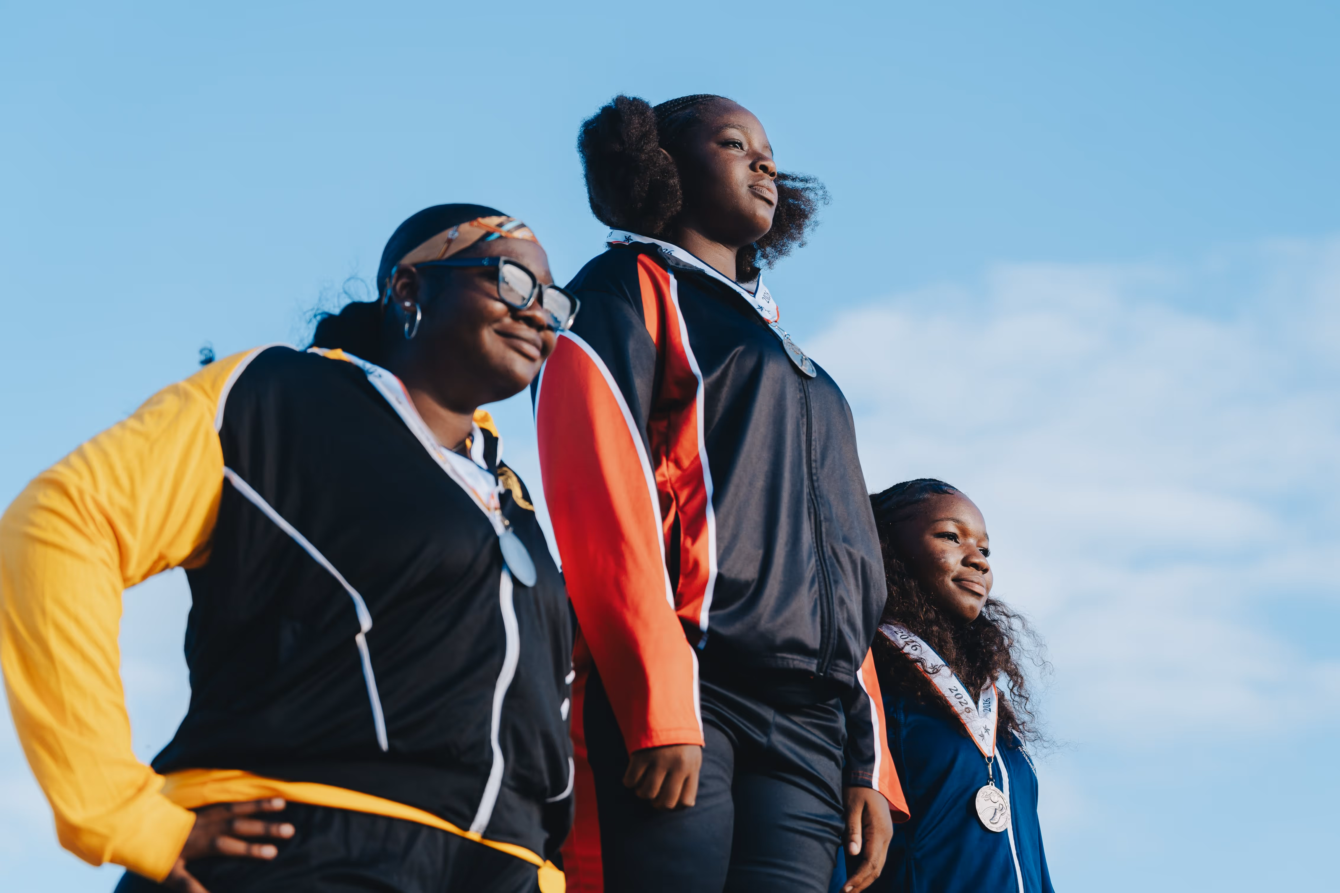 Three female athletes wearing medals and sports jackets stand confidently against a blue sky.