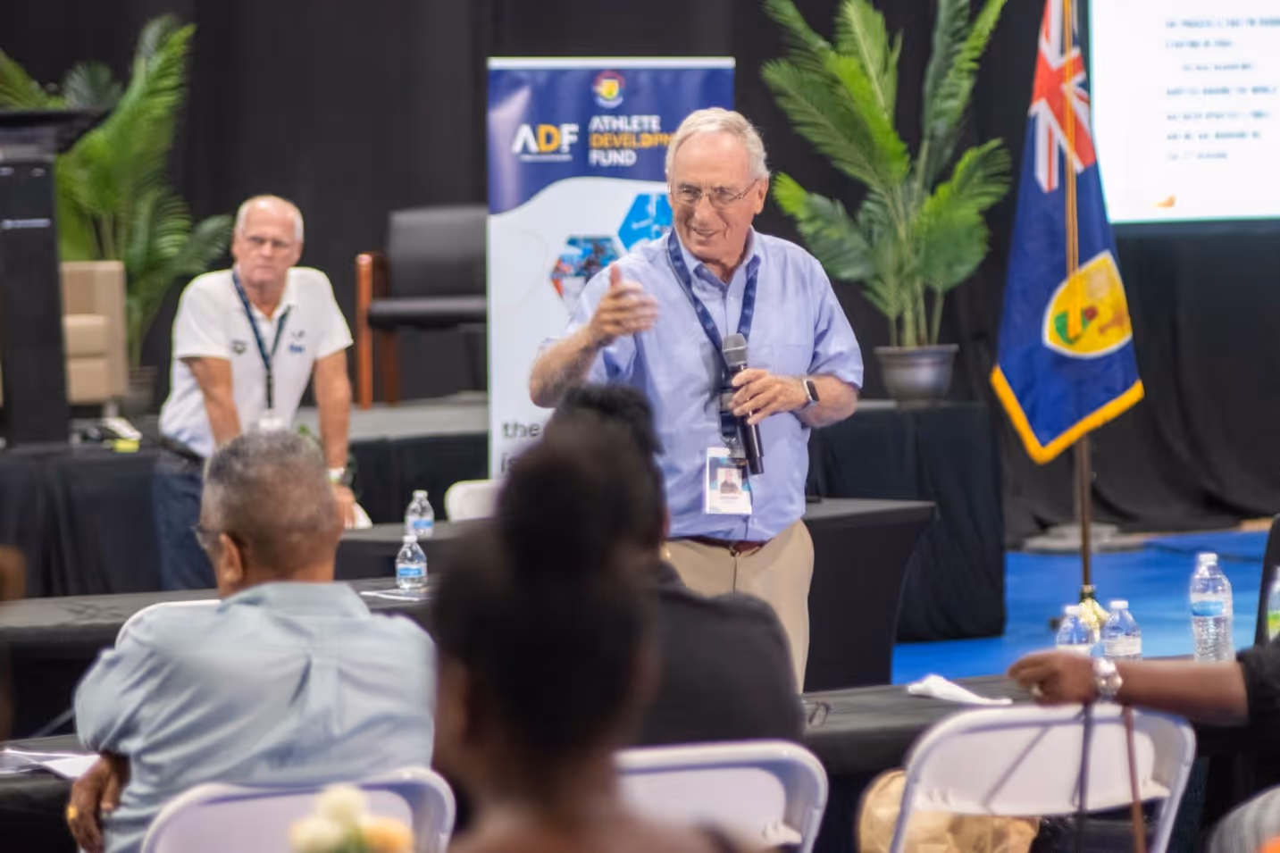 Older man with microphone speaking to seated audience at a conference with a flag and banner in background.