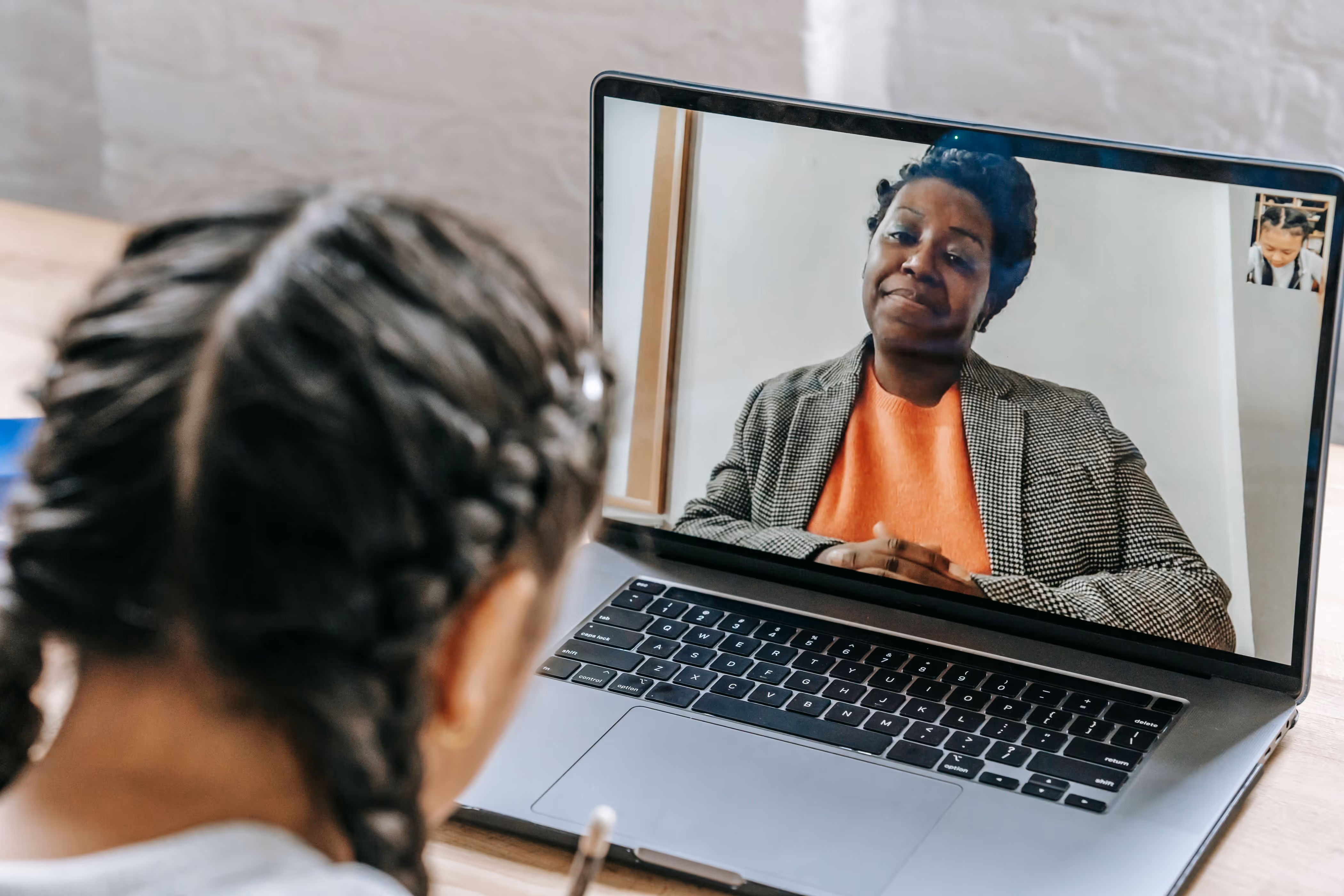 Child with braided hair attending a video call on a laptop with an adult woman in an orange shirt.