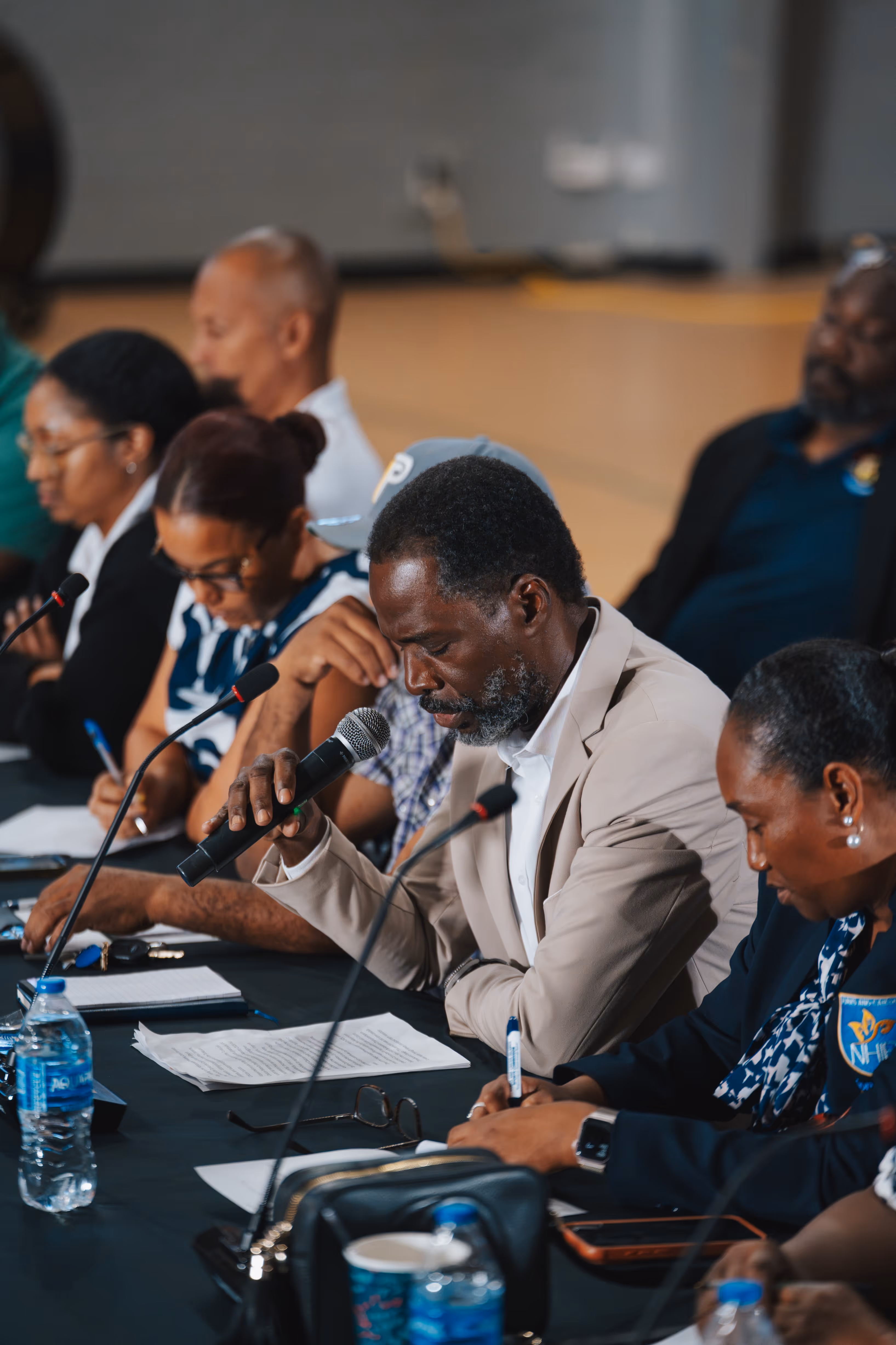 Man in beige jacket speaking into microphone while others sit and write at a conference table.