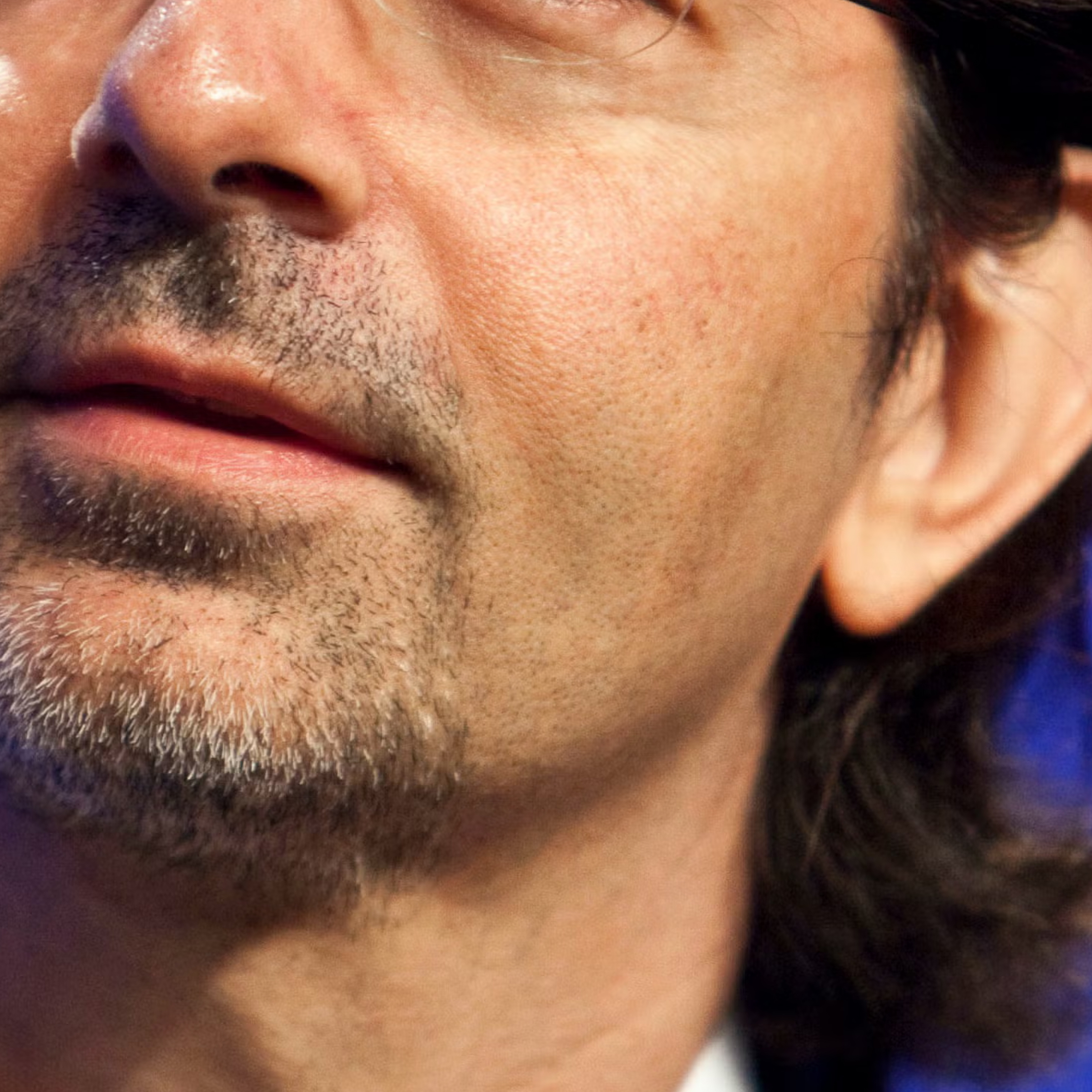 Close-up of a man's lower face with light stubble and long dark hair near his ear.