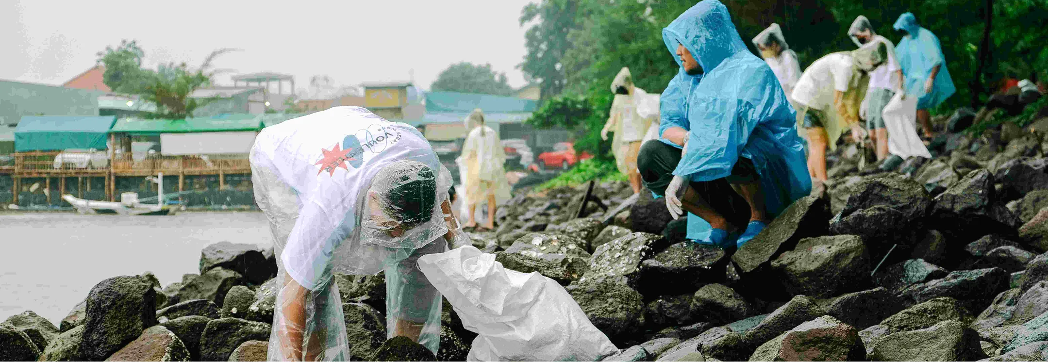 People wearing rain ponchos collecting trash on rocky shoreline near water on a rainy day.