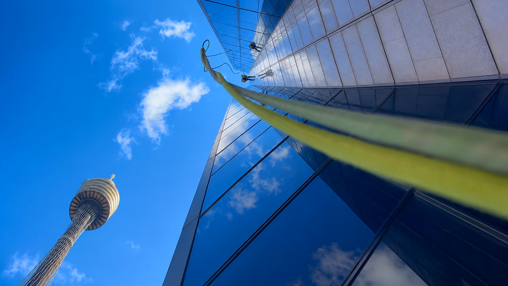 Upward view of a rope access technician descending a mirrored glass tower with Sydney Tower against blue sky