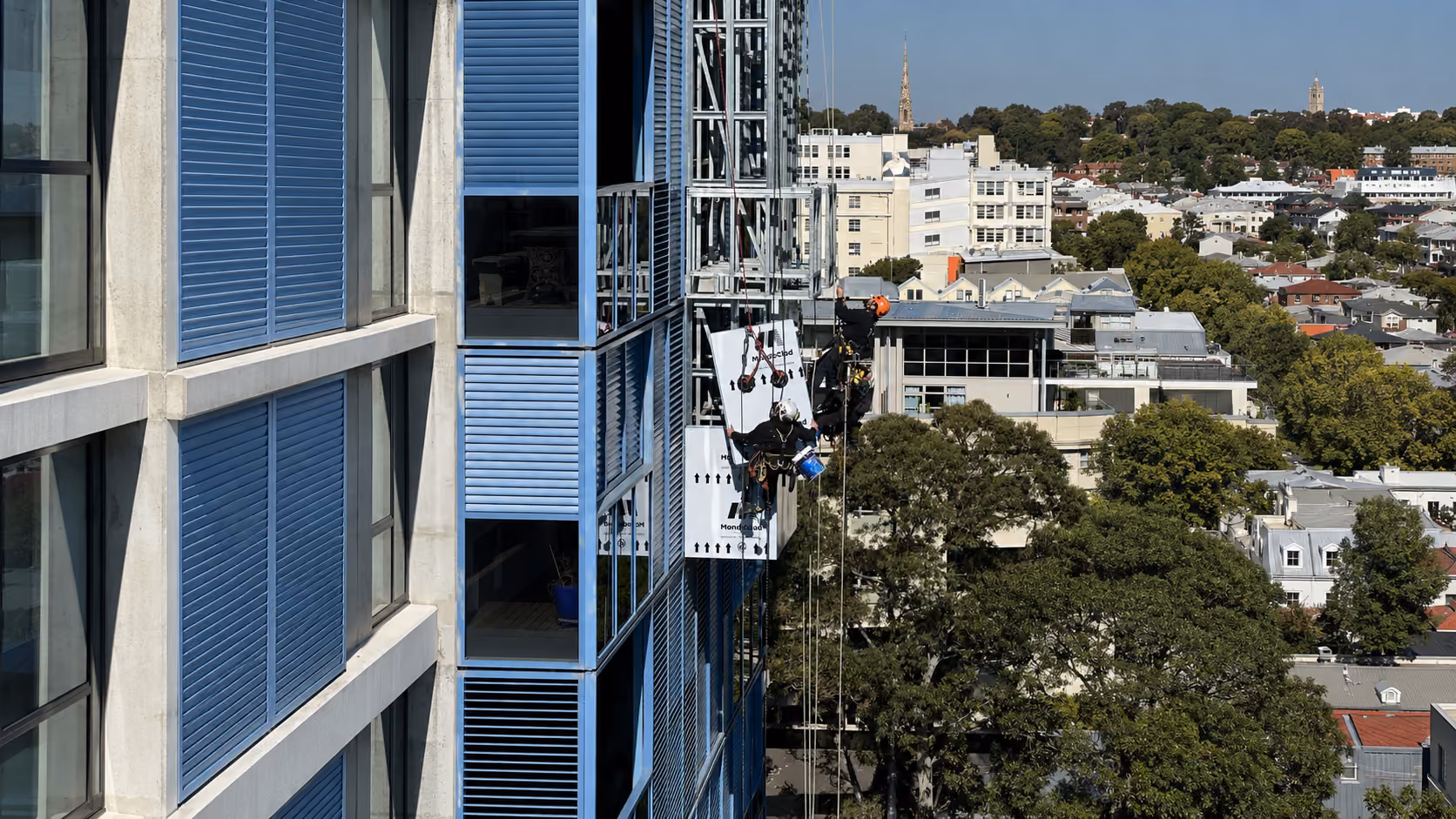 Two rope access technicians installing cladding panels on a high-rise facade above a leafy inner-suburban skyline
