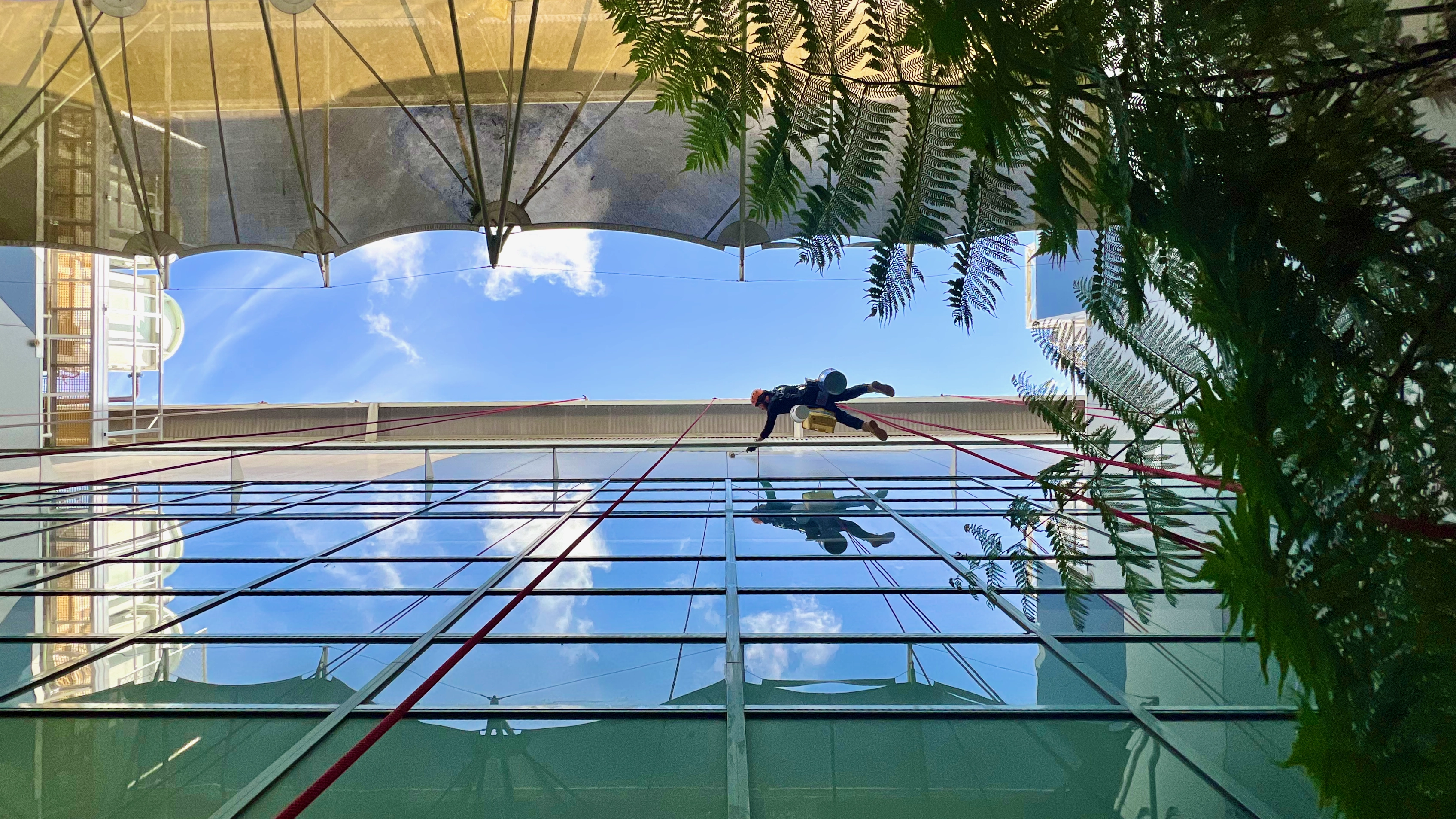 Looking up directly under a rope access technician who is extending out horizontally to reach an area of the facade that is being painted.