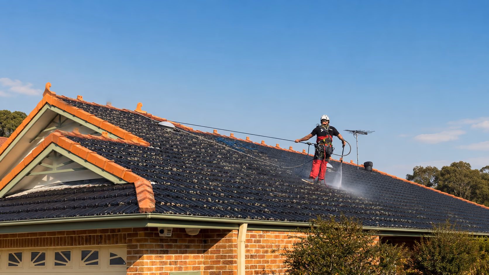 Harnessed technician pressure-washing a dark tiled pitched roof of a brick home under clear blue sky