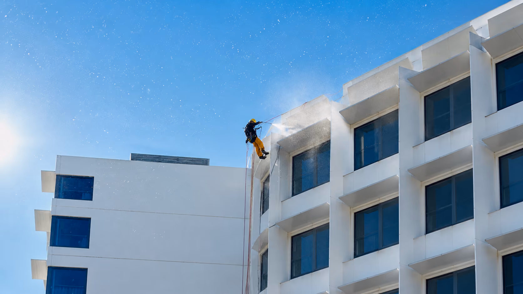 Rope access technician in yellow abseiling a white high-rise facade while pressure-washing, spray catching the sun