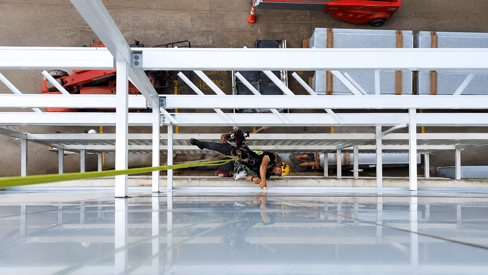 Rope access technician suspended horizontally beneath a white steel-framed structure, sealing the joints of a facade panel