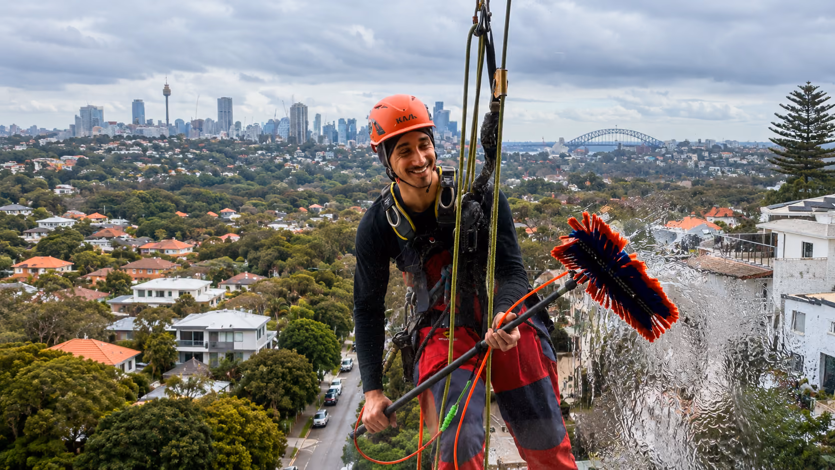 Smiling rope access window cleaner on ropes with squeegee pole, Sydney CBD skyline and Harbour Bridge behind