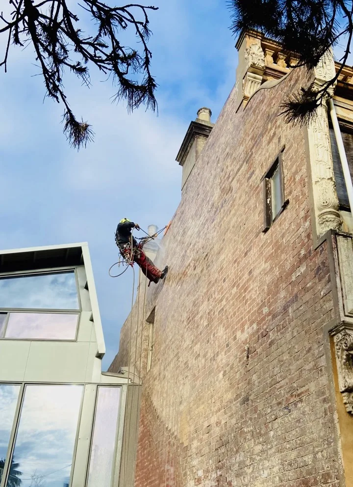 Rope-access technician soft-washing a heritage sandstone-trimmed brick facade in preparation for painting, Sydney