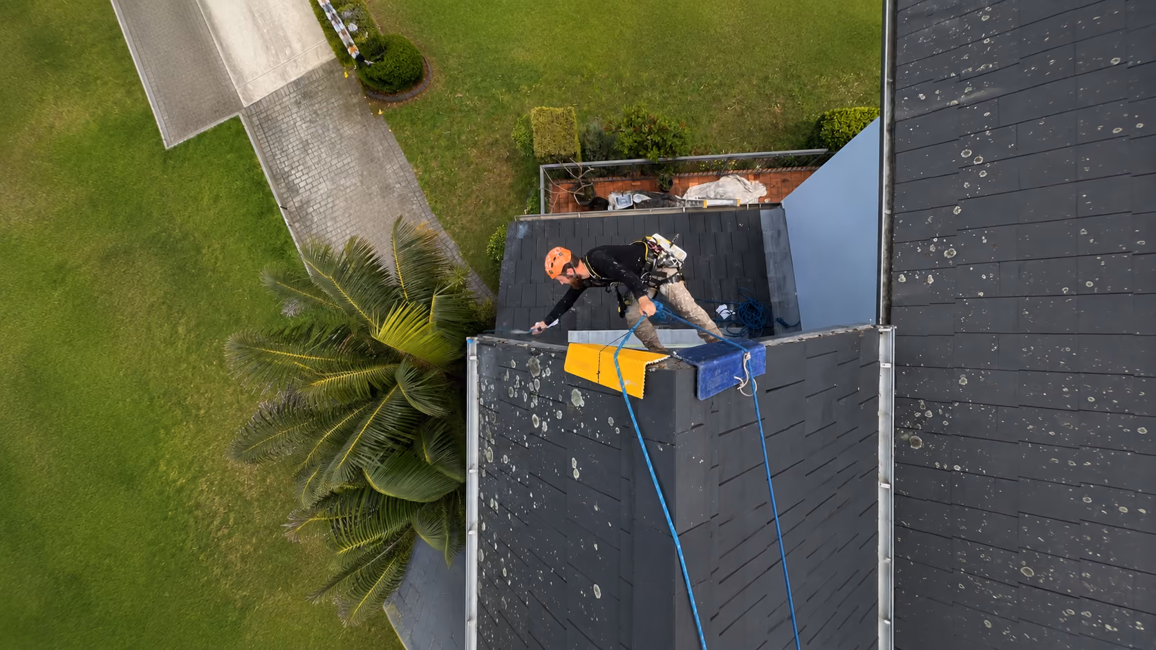 Aerial view of a harnessed rope access technician working along the ridge of a dark slate roof above a lawned garden painting the fascia boards