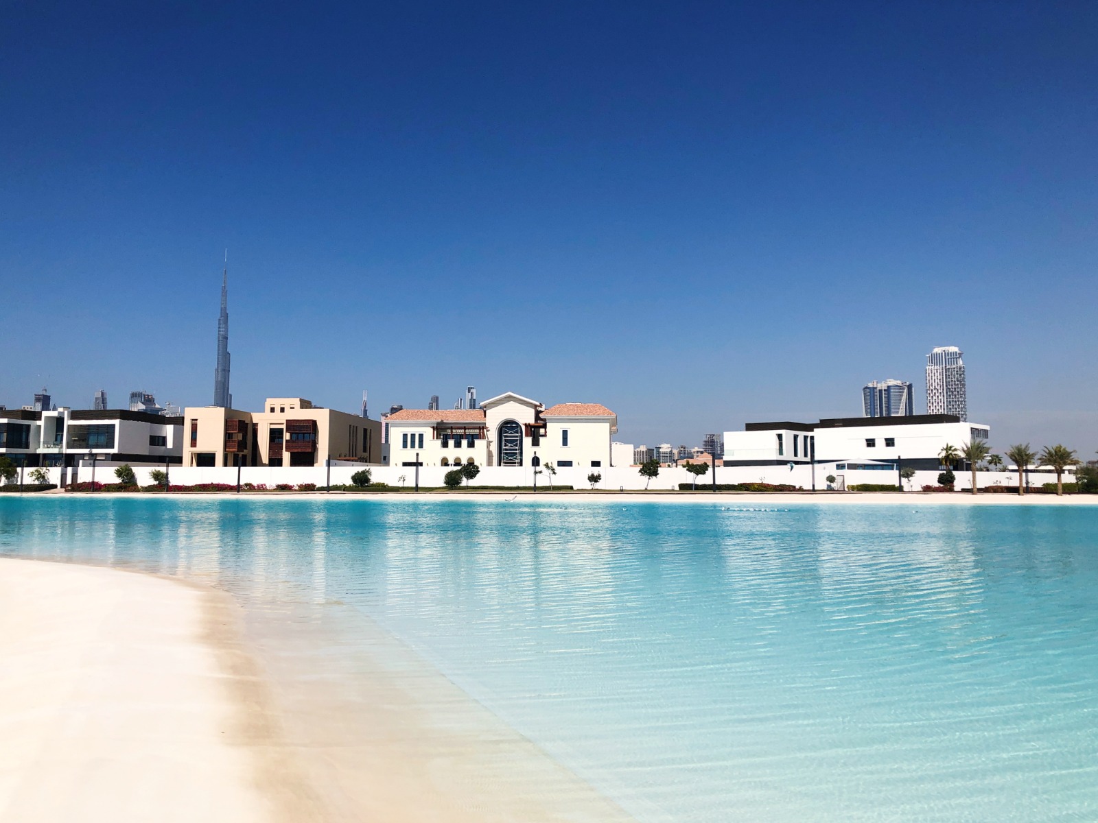Calm blue water in foreground with modern villas and Burj Khalifa in Dubai skyline under clear sky.