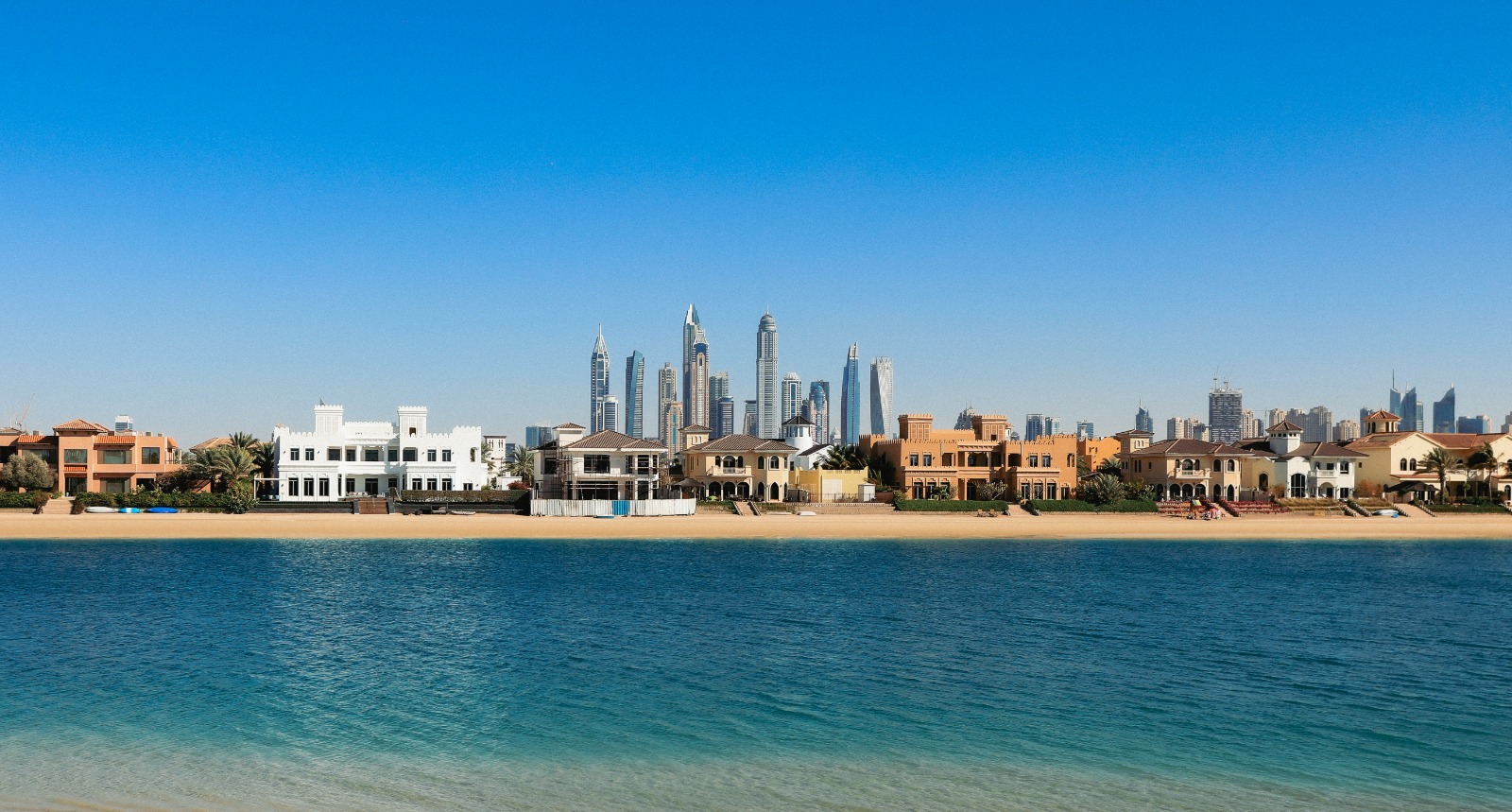 Waterfront villas on sandy shore with modern skyscrapers in the background under clear blue sky.