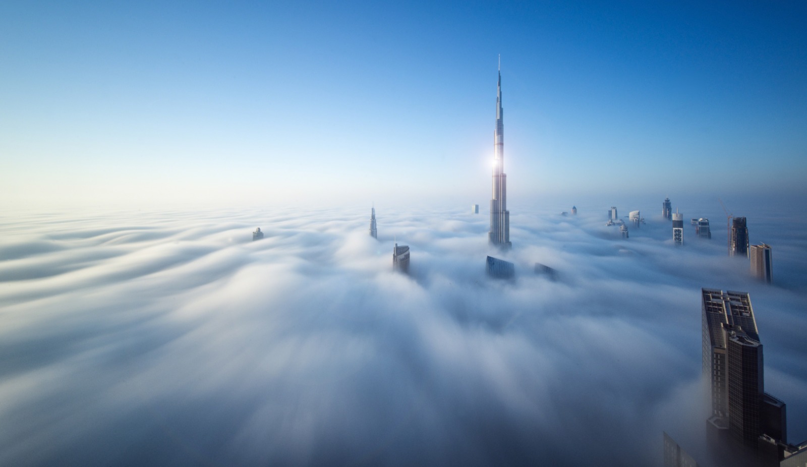 Skyscrapers, including Burj Khalifa, rising above a thick fog under a clear blue sky.