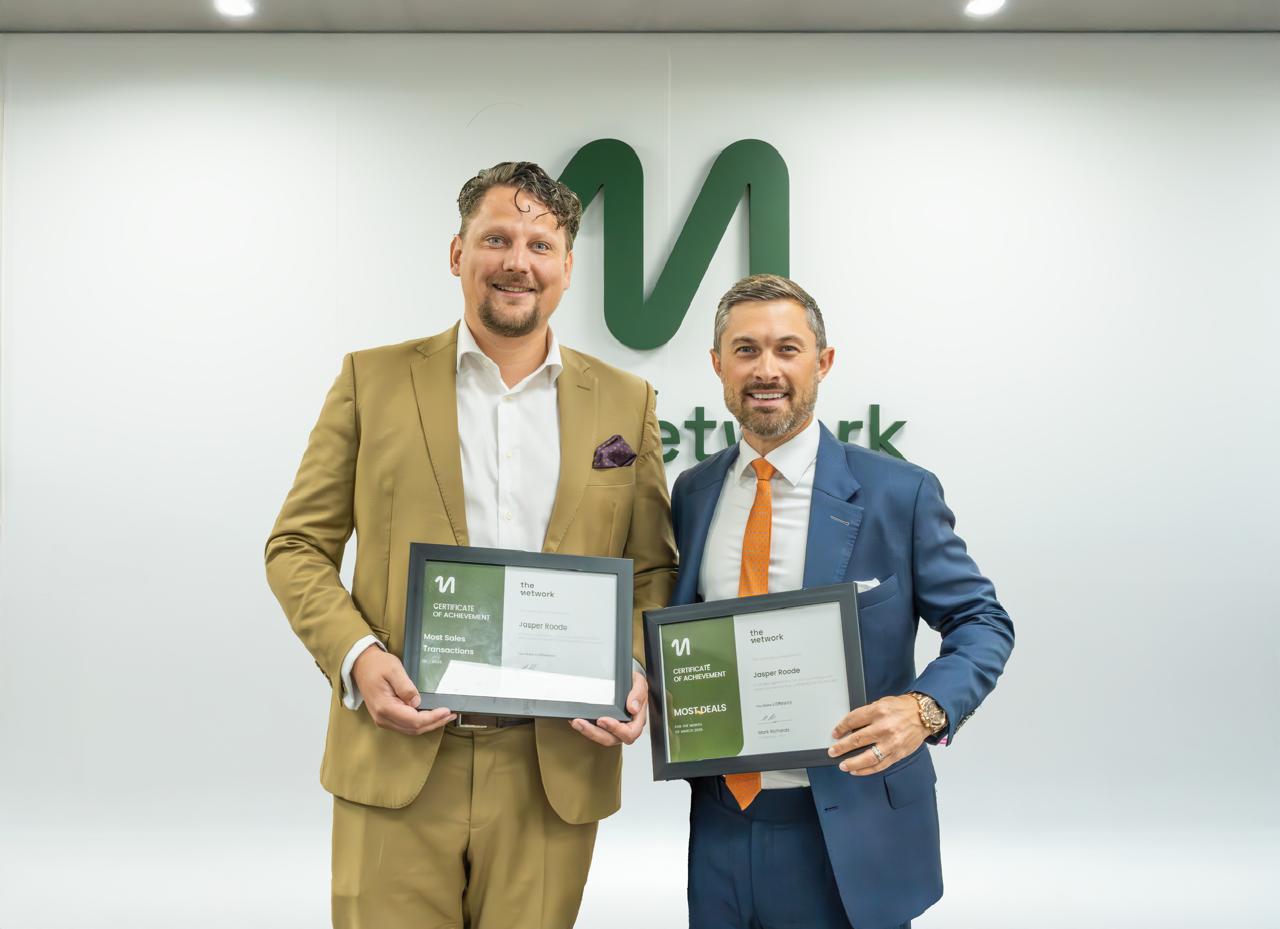 Two men in suits smiling, each holding a framed certificate of achievement in front of a white wall with logo.