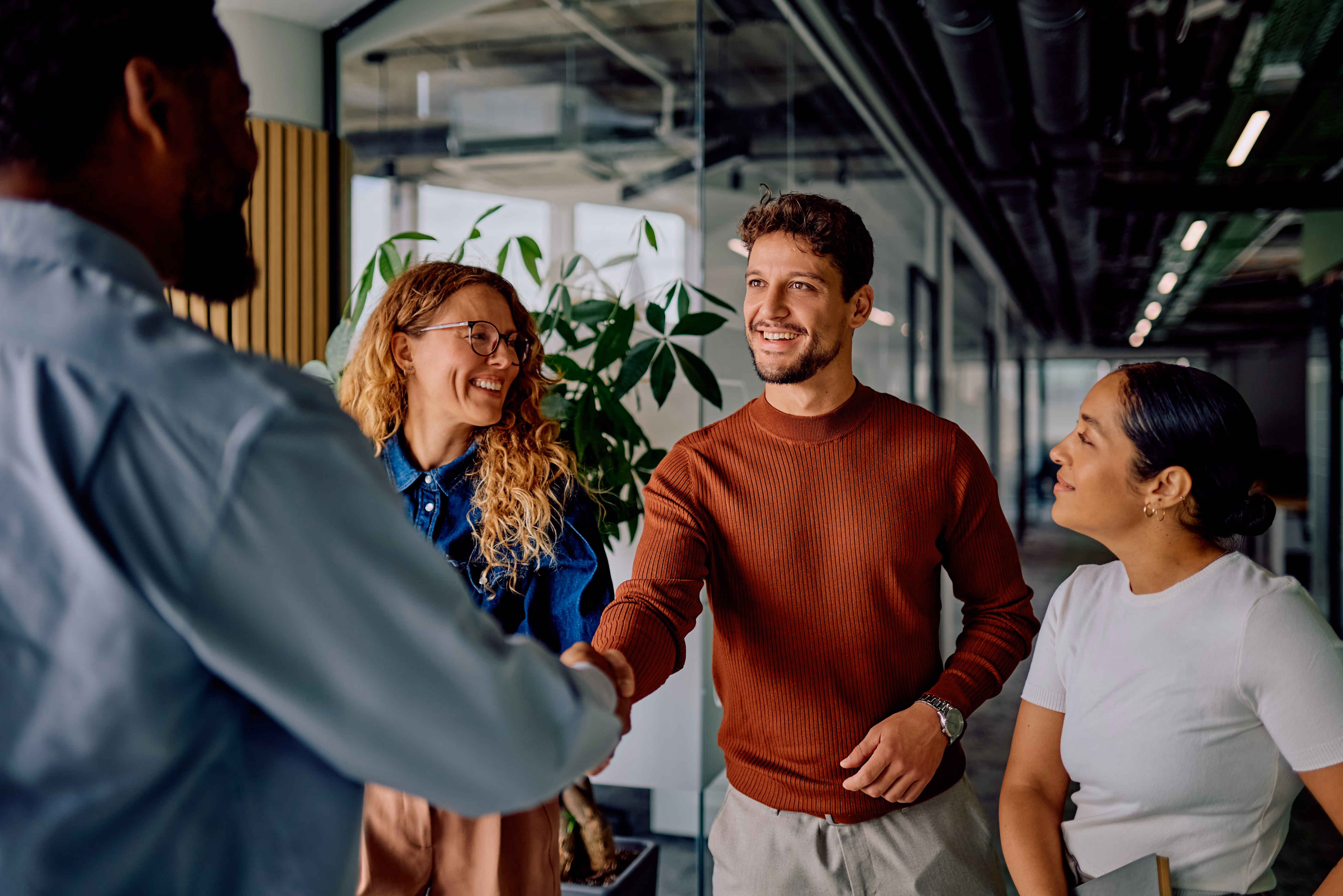 Man with brown sweater shaking hands with another man while two women watch and smile in office.