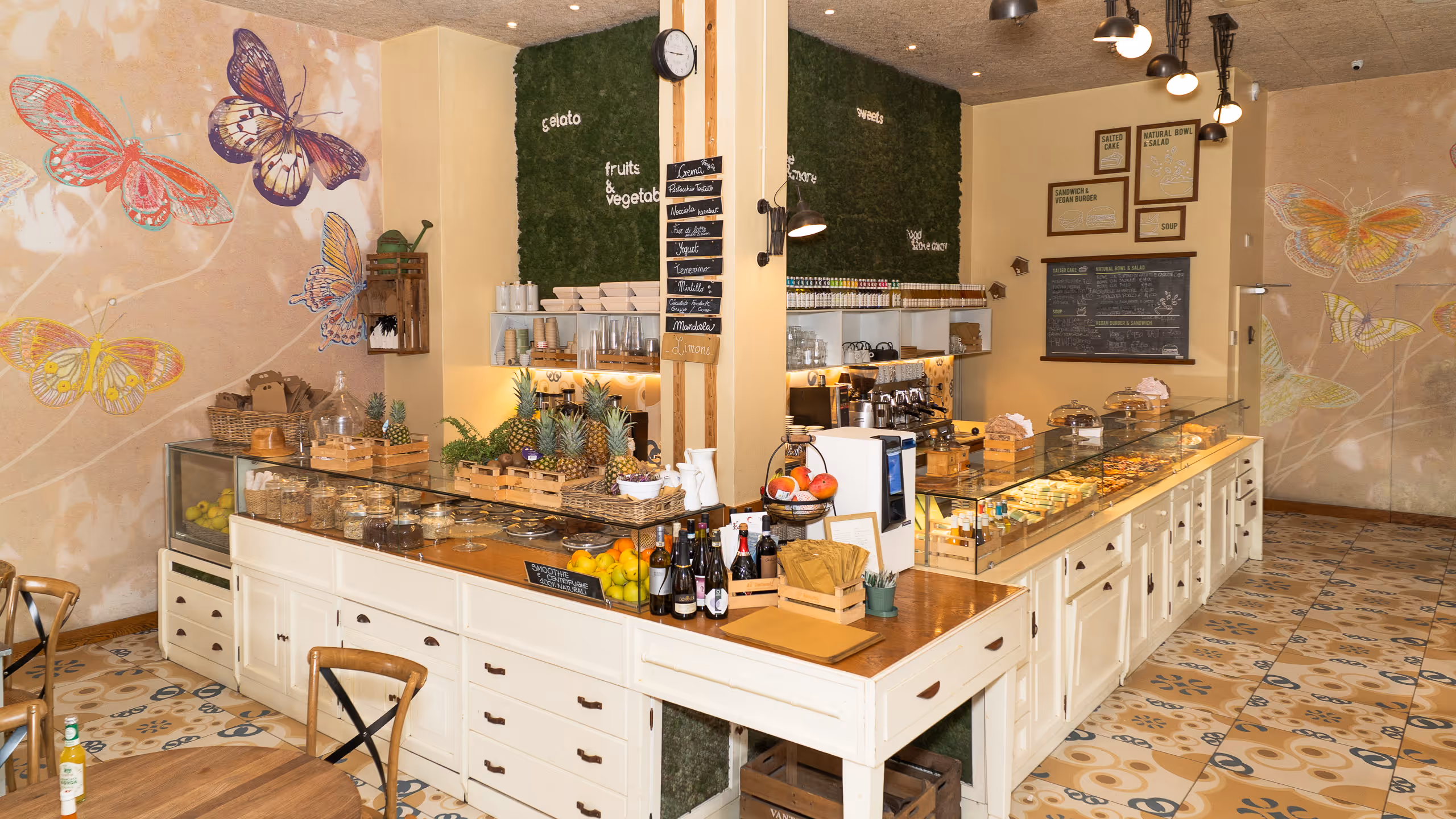Interno di un negozio con banco in legno, frutta, bottiglie e muro decorato con farfalle colorate.
