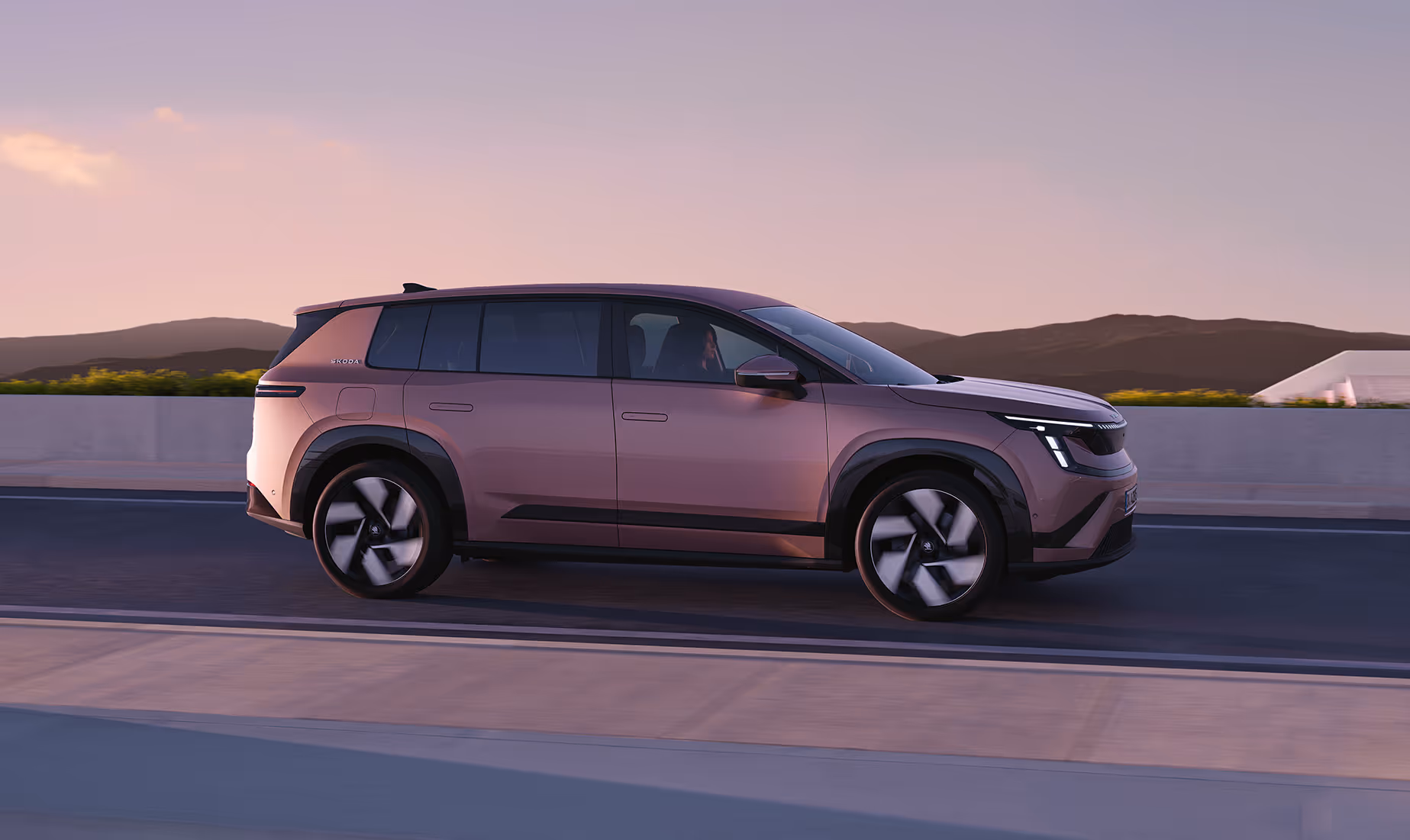 Dynamische Seitenansicht des neuen Škoda Peaq in der Farbe Rosewood-Beige Metallic während der Fahrt auf einer Straße vor hügeliger Kulisse im sanften Abendlicht. Das modern gezeichnete SUV zeichnet sich durch eine kantige Silhouette, flächenbündige Türgriffe und markant abgesetzte, dunkle Radkästen aus.