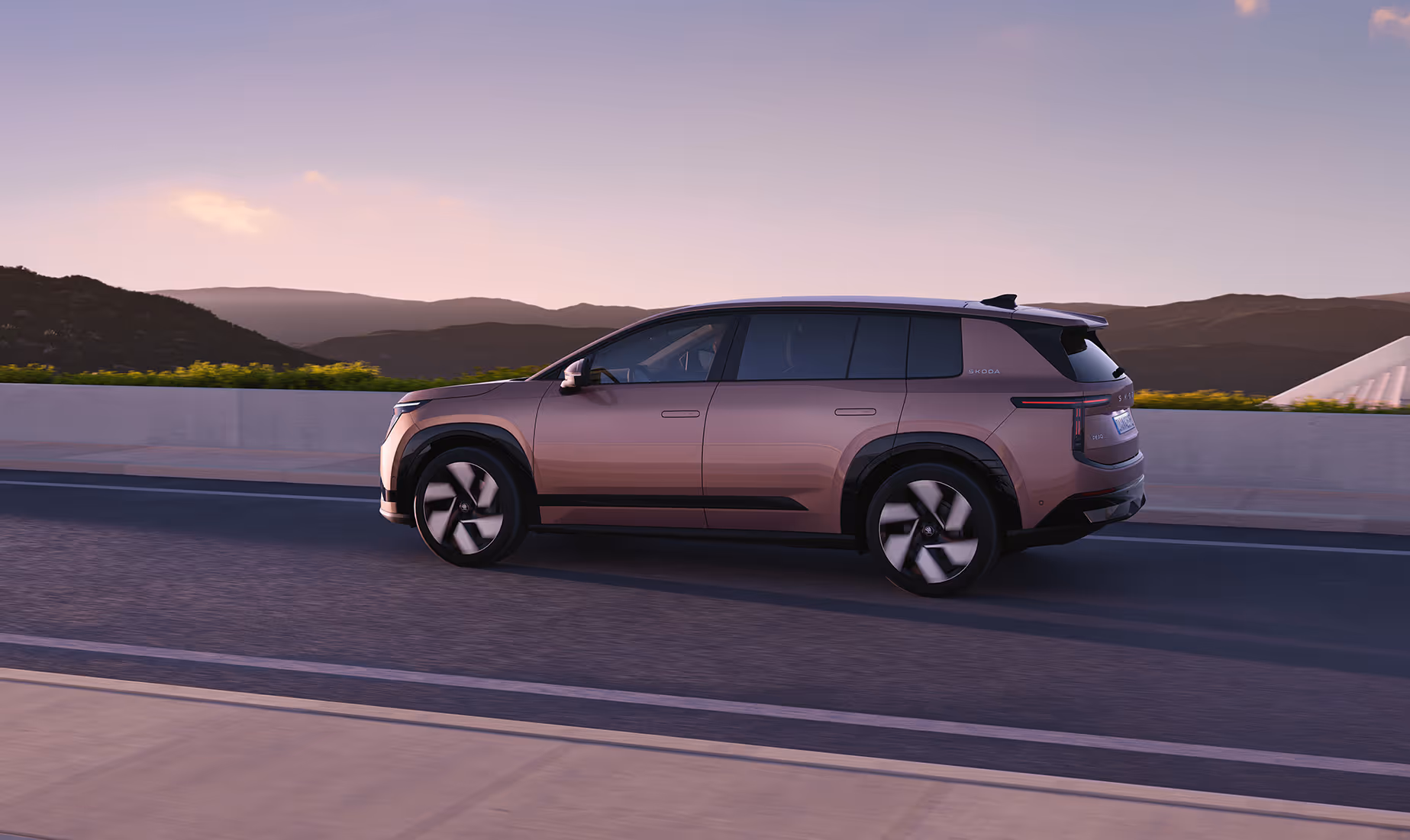 Dynamische Heck- und Seitenansicht des Škoda Peaq in der Farbe Rosewood-Beige Metallic während der Fahrt auf einer Straße im sanften Abendlicht. Die klare Linienführung des SUVs wird durch flächenbündige Türgriffe, dunkel abgesetzte Radhäuser und eine markante, modern gestaltete Heckpartie betont. Drehende, aerodynamische Leichtmetallfelgen verleihen dem Fahrzeug im Zusammenspiel mit der hügeligen Landschaftskulisse zusätzliche Dynamik.