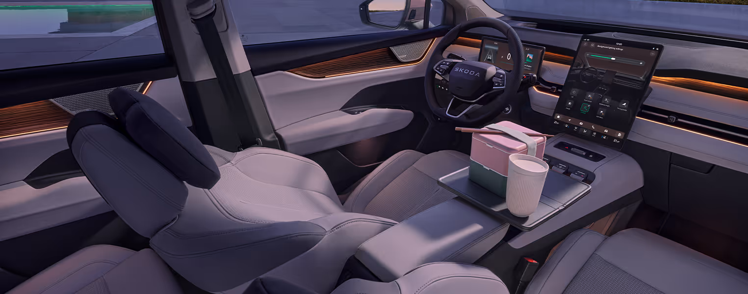 Blick in das vordere Interieur des Škoda Peaq mit keramikfarbener Ausstattung, bei dem der Fahrersitz weit in eine entspannte Ruheposition zurückgeneigt ist. Über der Mittelkonsole ist ein kleiner Tisch ausgeklappt, auf dem eine gestapelte Lunchbox mit Essstäbchen und ein Becher abgestellt sind. Im Hintergrund komplettieren das Multifunktionslenkrad, ein großes, hochformatiges Infotainment-Display und die warme Ambientebeleuchtung das moderne Cockpit.
