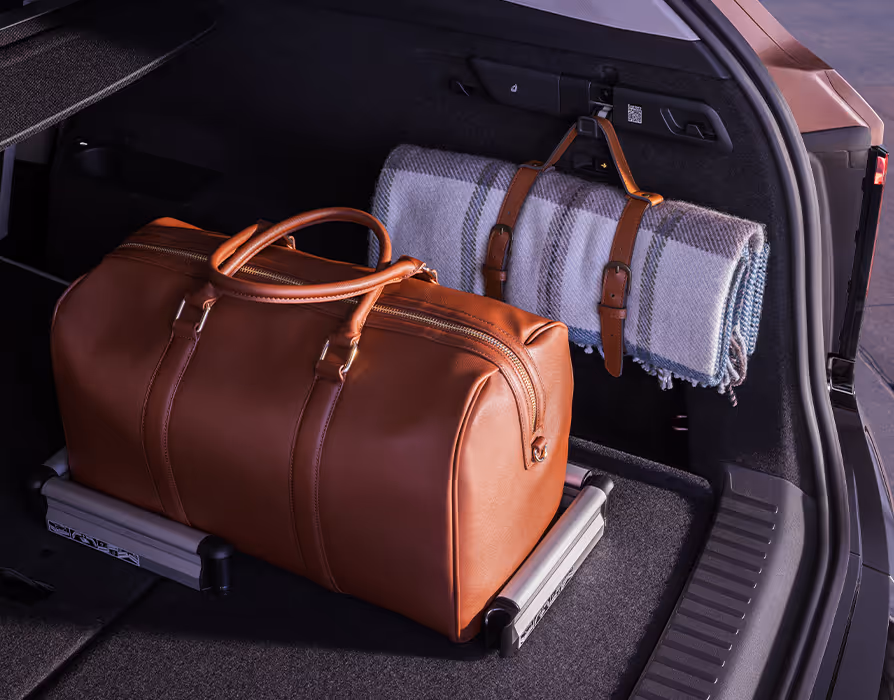 Blick in den rechten Bereich des geöffneten Kofferraums im Škoda Peaq. Auf dem dunklen Ladeboden steht eine braune Reisetasche, die durch Cargoelemente sicher gegen Verrutschen fixiert ist. An der Seitenverkleidung hängt zudem eine aufgerollte Decke mit Lederriemen griffbereit an einem integrierten Taschenhaken.