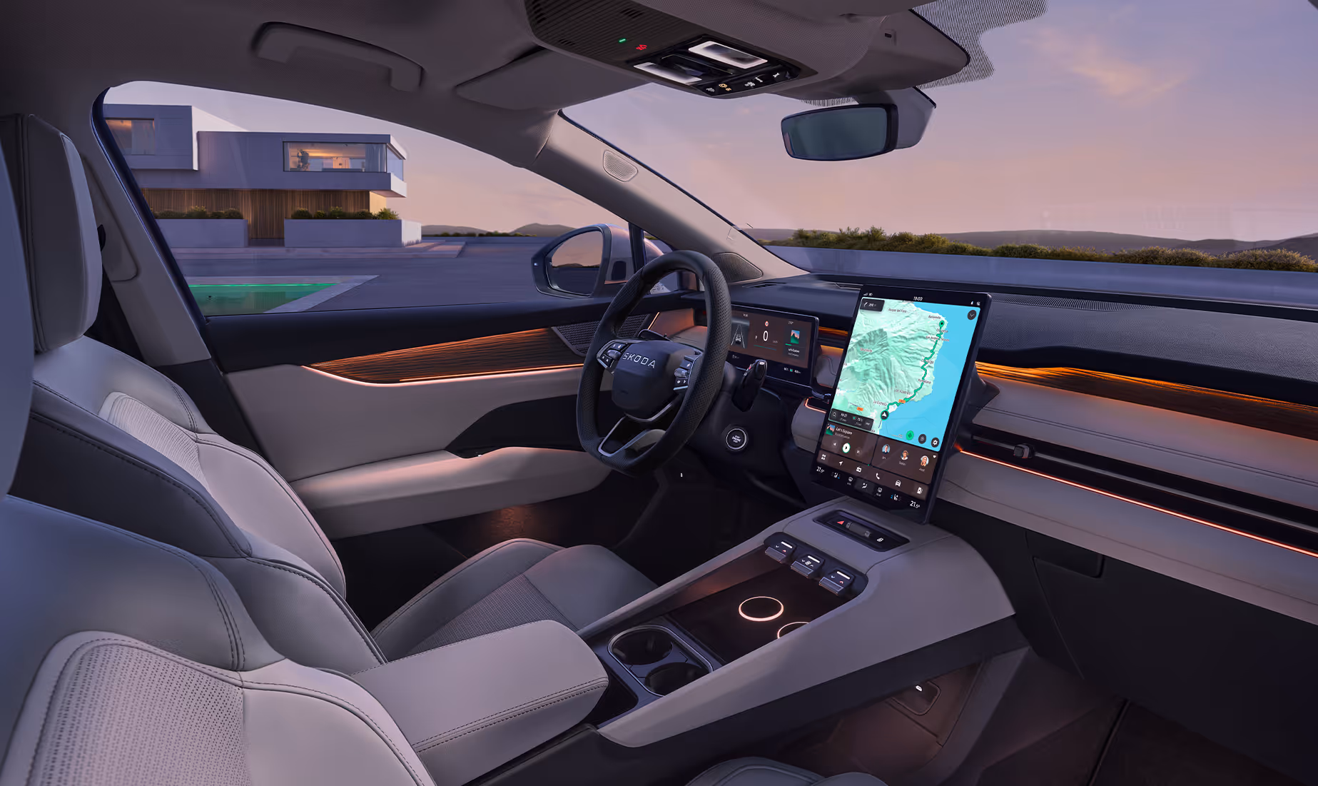 Blick von der Beifahrerseite in das aufgeräumte Cockpit des Škoda Peaq, dessen helle keramikfarbene Ausstattung durch eine warme Ambientebeleuchtung dezent in Szene gesetzt wird. Im Zentrum der Instrumententafel dominiert ein großes, hochformatiges Infotainment-Display mit aktiver Kartennavigation neben dem Multifunktionslenkrad. Durch die Fensterscheiben fällt der Blick im sanften Abendlicht auf eine moderne Villa mit Pool.