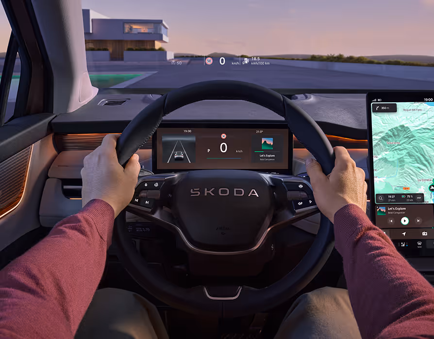Blick aus der Fahrerperspektive in das moderne Cockpit des Škoda Peaq bei abendlichen Lichtverhältnissen. Die Hände ruhen auf dem Multifunktionslenkrad, hinter dem ein digitales Kombiinstrument sowie ein Head-up-Display auf der Windschutzscheibe wichtige Fahrdaten anzeigen. Rechts daneben ist ein großes, freistehendes Infotainment-Display mit aktiver Routenführung zu sehen, während eine warme Ambientebeleuchtung das Interieur dezent akzentuiert.