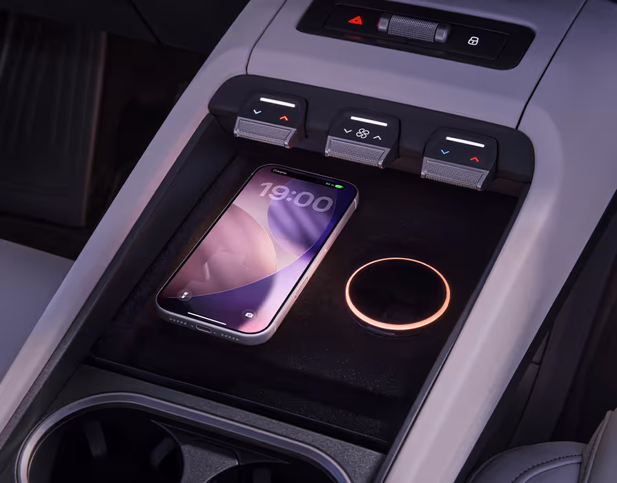 Detailaufnahme der vorderen Mittelkonsole im Škoda Peaq. Auf der dunklen Ablagefläche liegt ein Smartphone auf einer induktiven Ladestation, deren Ladebereich durch einen warm leuchtenden Ring optisch hervorgehoben wird. Direkt darüber befinden sich drei haptische Drehregler zur Fahrzeugbedienung, während im unteren Bildbereich zwei integrierte Getränkehalter zu erkennen sind.