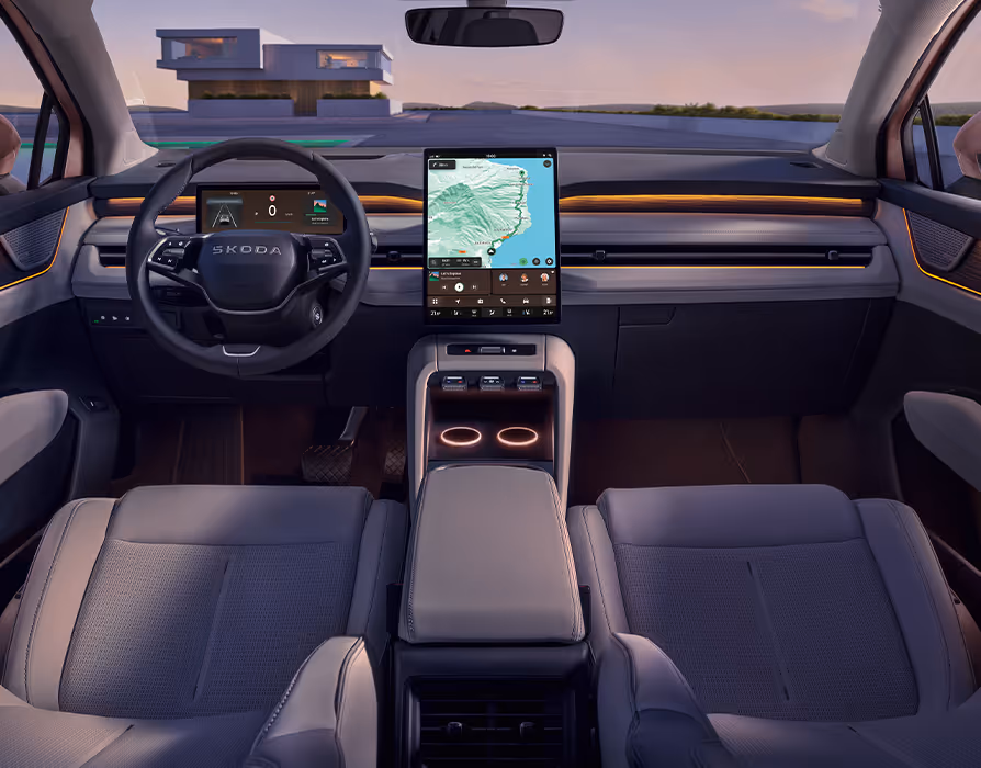 Symmetrischer Blick aus dem Fond in das Cockpit des Škoda Peaq mit der hellen Innenausstattung "Design Selection Suite Keramik". Das aufgeräumte Interieur wird von keramikfarbenen Vordersitzen, einem Multifunktionslenkrad und einem großen, zentralen Infotainment-Display mit aktiver Navigation geprägt. Eine warme, orangefarbene Ambientebeleuchtung entlang der Armaturentafel und in der Mittelkonsole sorgt im abendlichen Licht für eine stimmungsvolle Atmosphäre.