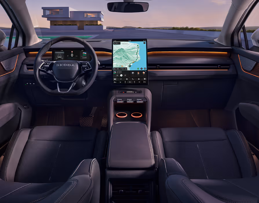 Symmetrischer Blick aus dem Fond in das Cockpit des Škoda Peaq mit der dunklen Innenausstattung "Design Selection Suite Schwarz". Das aufgeräumte Interieur wird von schwarzen Sitzen, dem Multifunktionslenkrad und einem großen, zentralen Infotainment-Display mit aktiver Navigation geprägt. Eine warme, orangefarbene Ambientebeleuchtung entlang der Armaturentafel und in der Mittelkonsole setzt im abendlichen Licht stimmungsvolle Akzente.